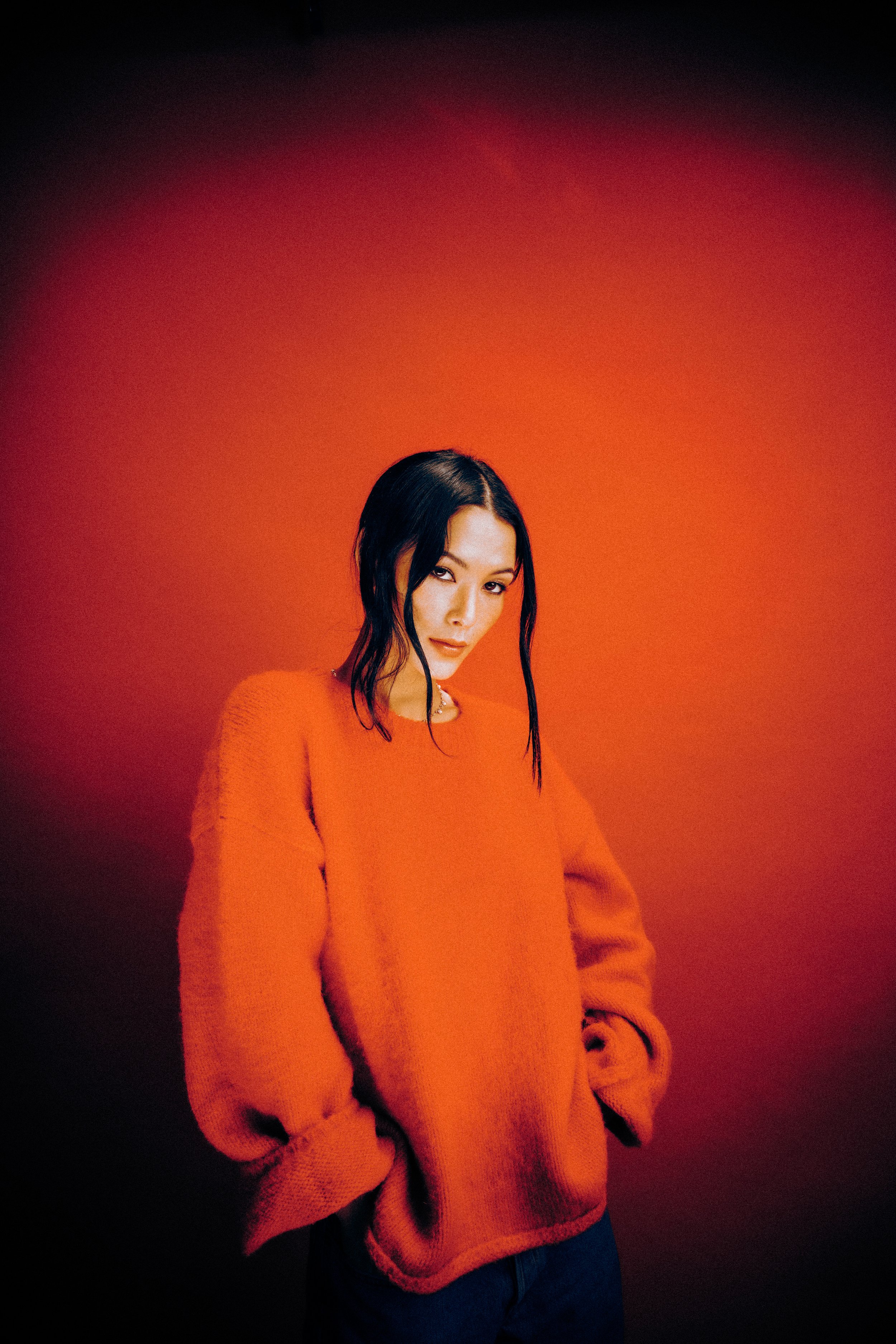 A woman with black hair wearing an orange sweater standing against an orange gradient background.