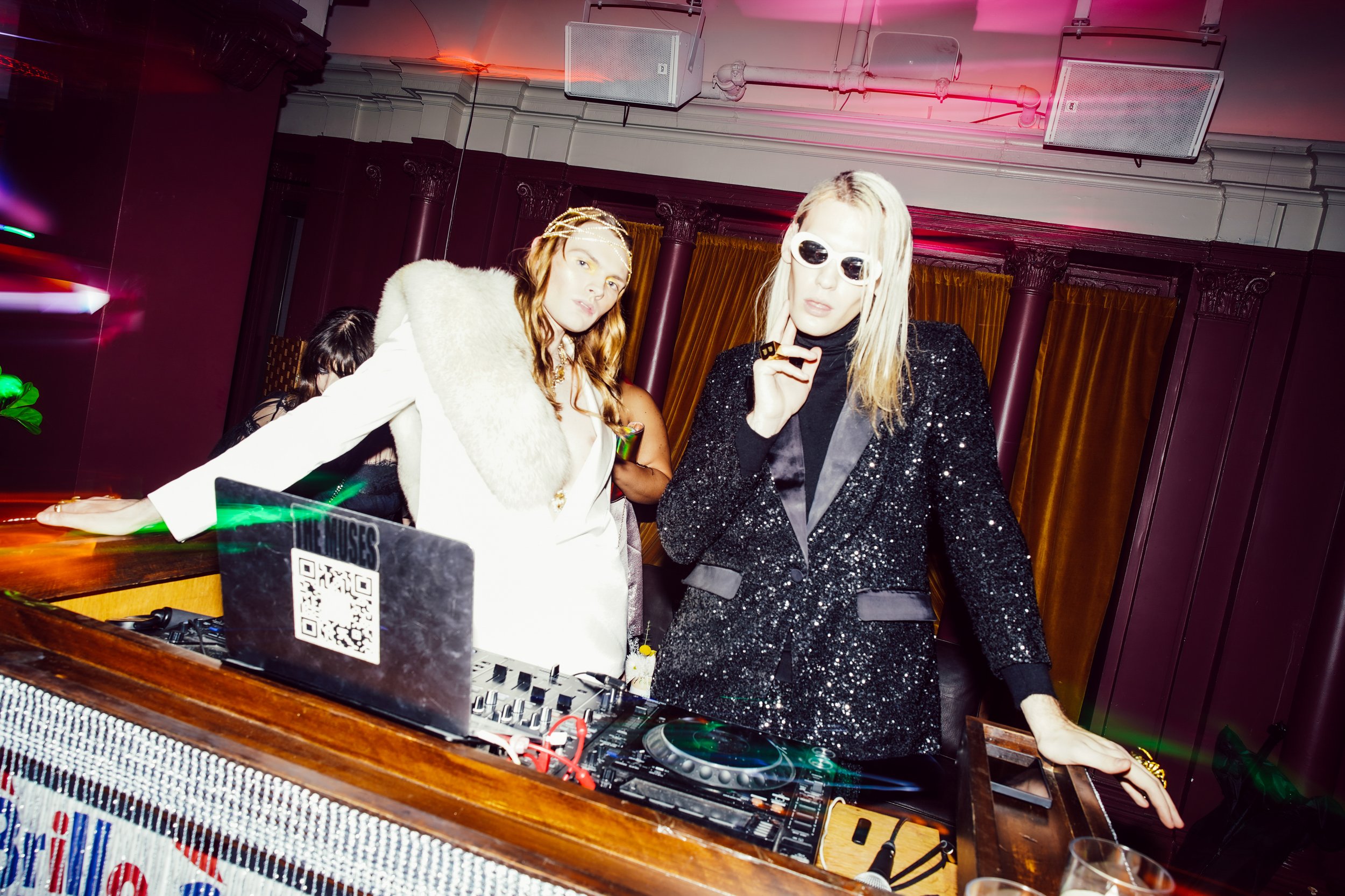 Two women dressed in glamorous outfits, one in a white coat with a fur collar and the other in a black sequined blazer, stand behind a DJ booth at a party. The woman in black is wearing white sunglasses and has platinum blonde hair, while the other w