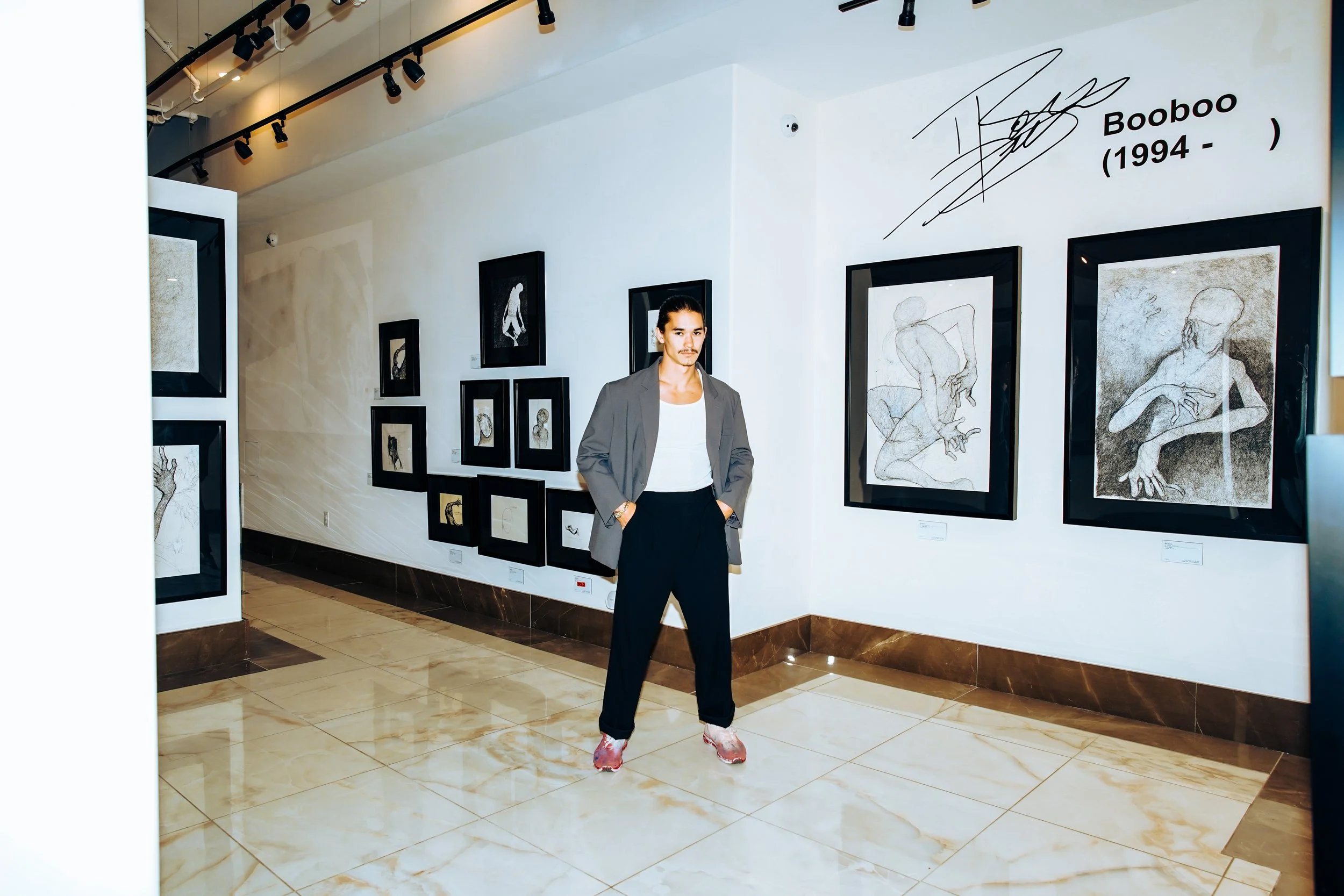 A man with a mustache and long hair tied back, wearing a gray blazer, white shirt, black pants, and plastic shoe covers, standing in an art gallery with framed drawings on the wall, including depictions of human figures, and a signature with the name