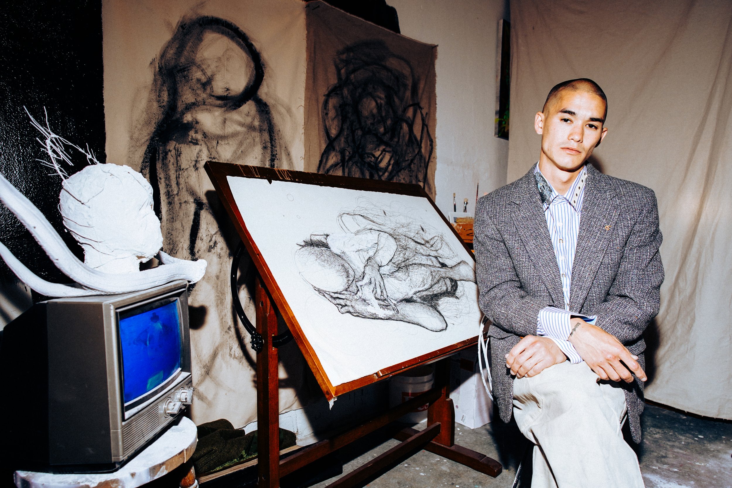 An artist posing in a studio with art supplies and sketches, wearing a blazer and light-colored pants.