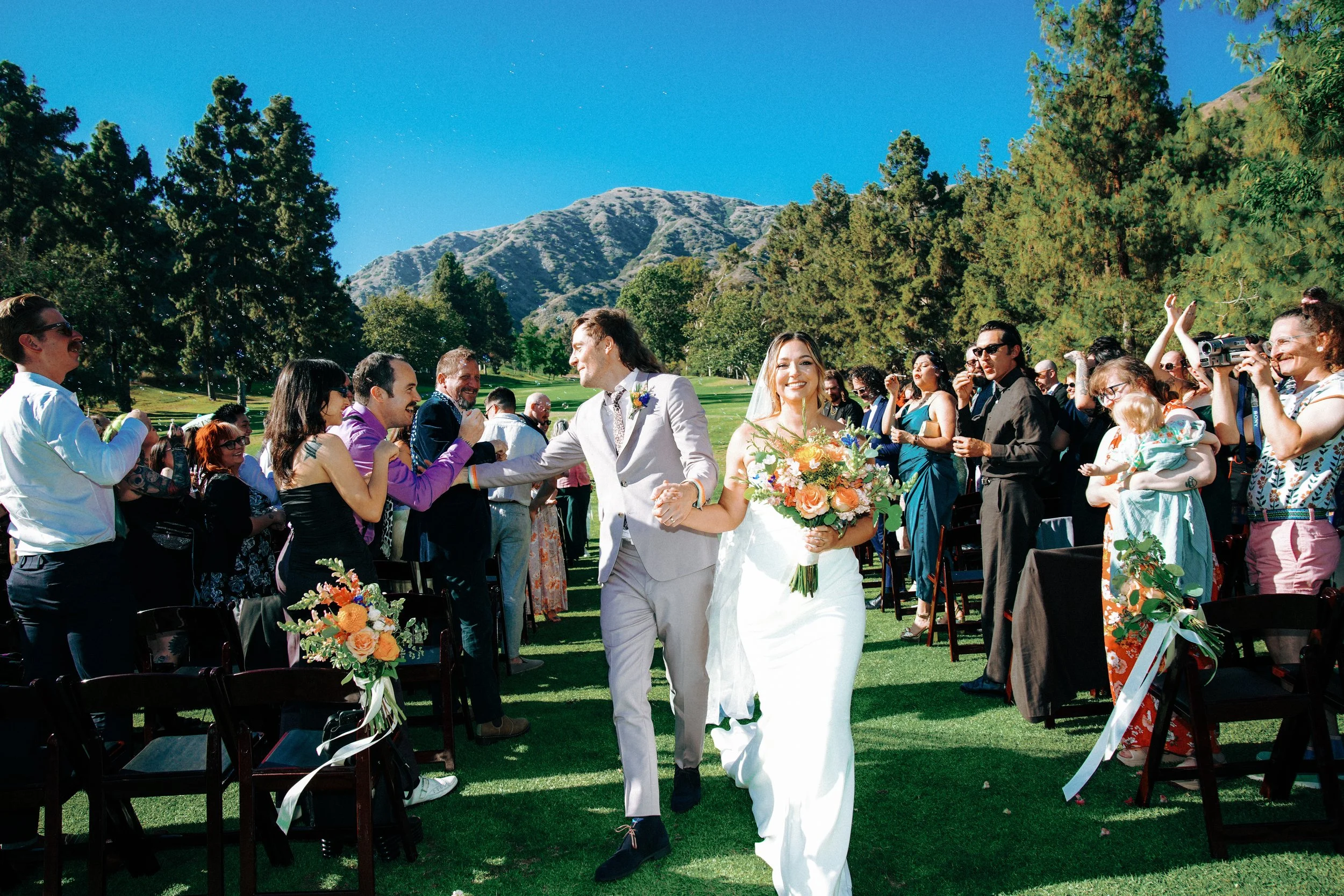 The image shows a newly married couple walking down the aisle with guests clapping and taking photos on a sunny outdoor wedding ceremony in a scenic mountain setting.