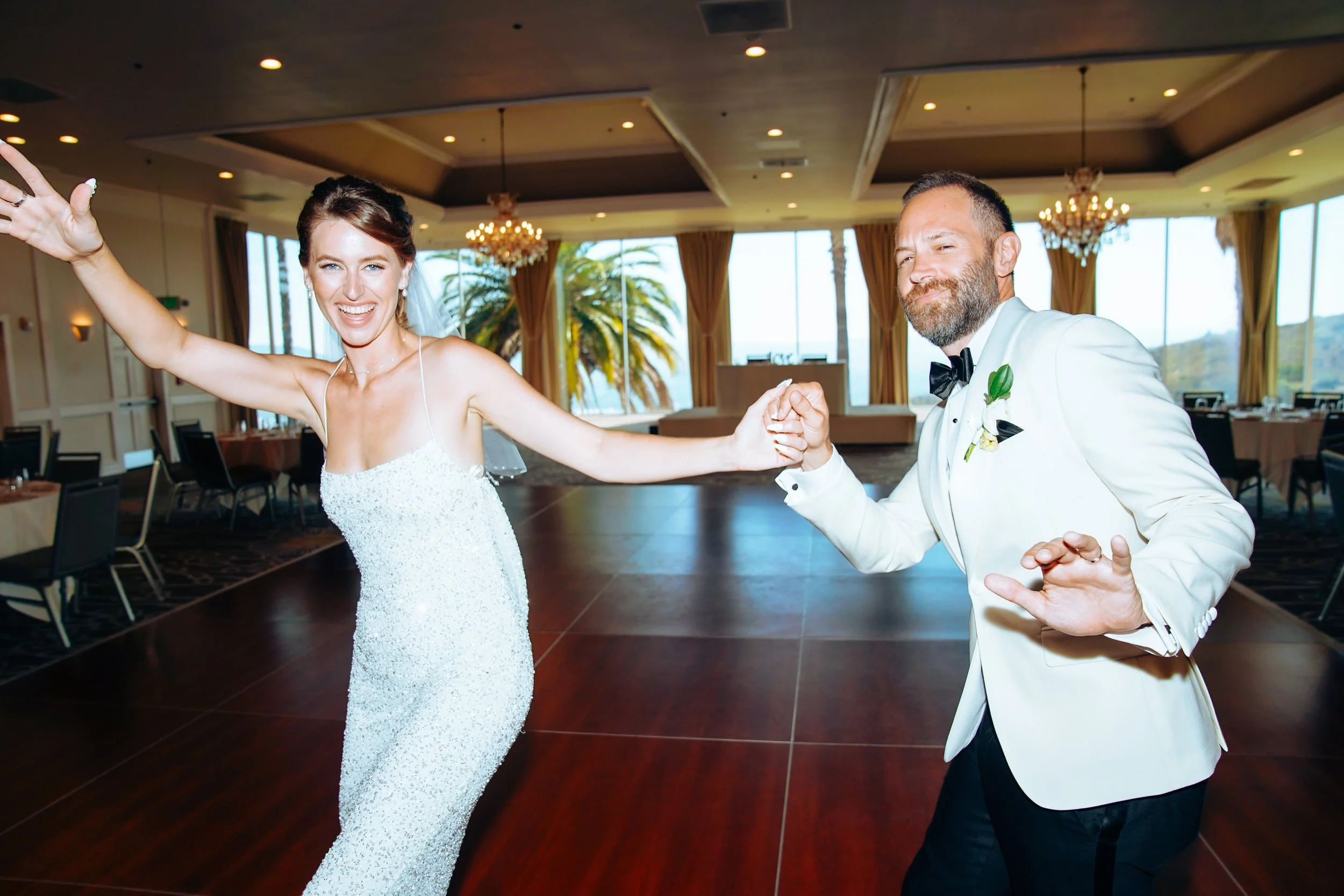 A bride and groom dancing at their wedding reception in a decorated indoor venue, with the bride in a white wedding gown and the groom in a white tuxedo with a black bow tie.