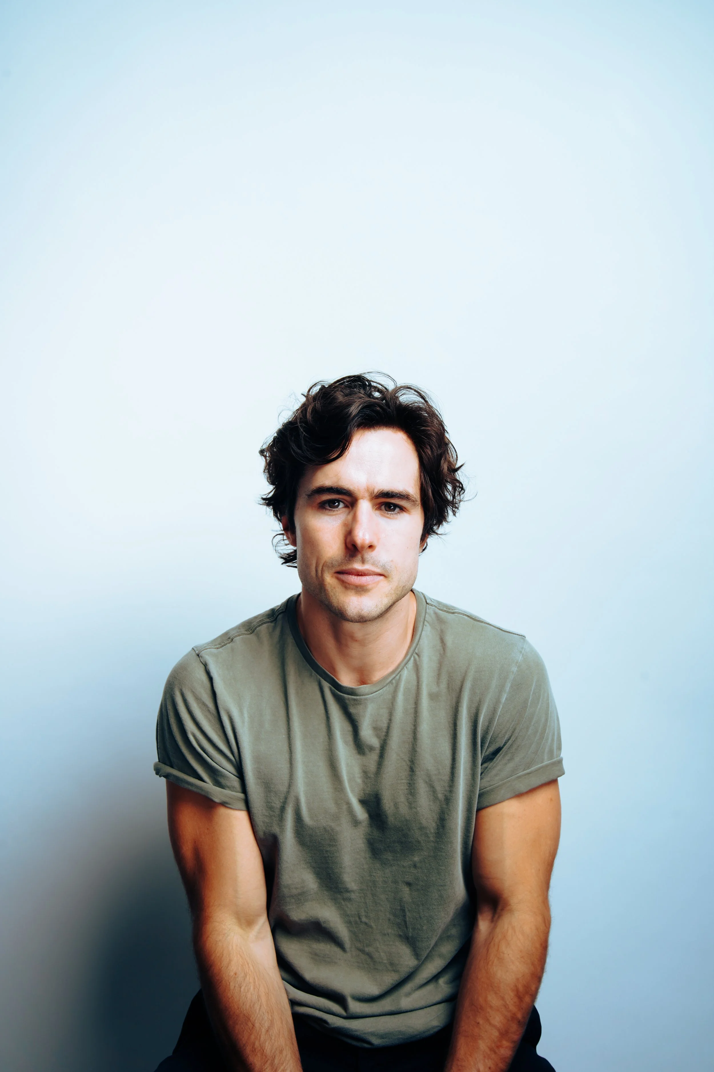 A portrait of a young man with dark, wavy hair, wearing a green t-shirt, looking directly at the camera against a light blue background.