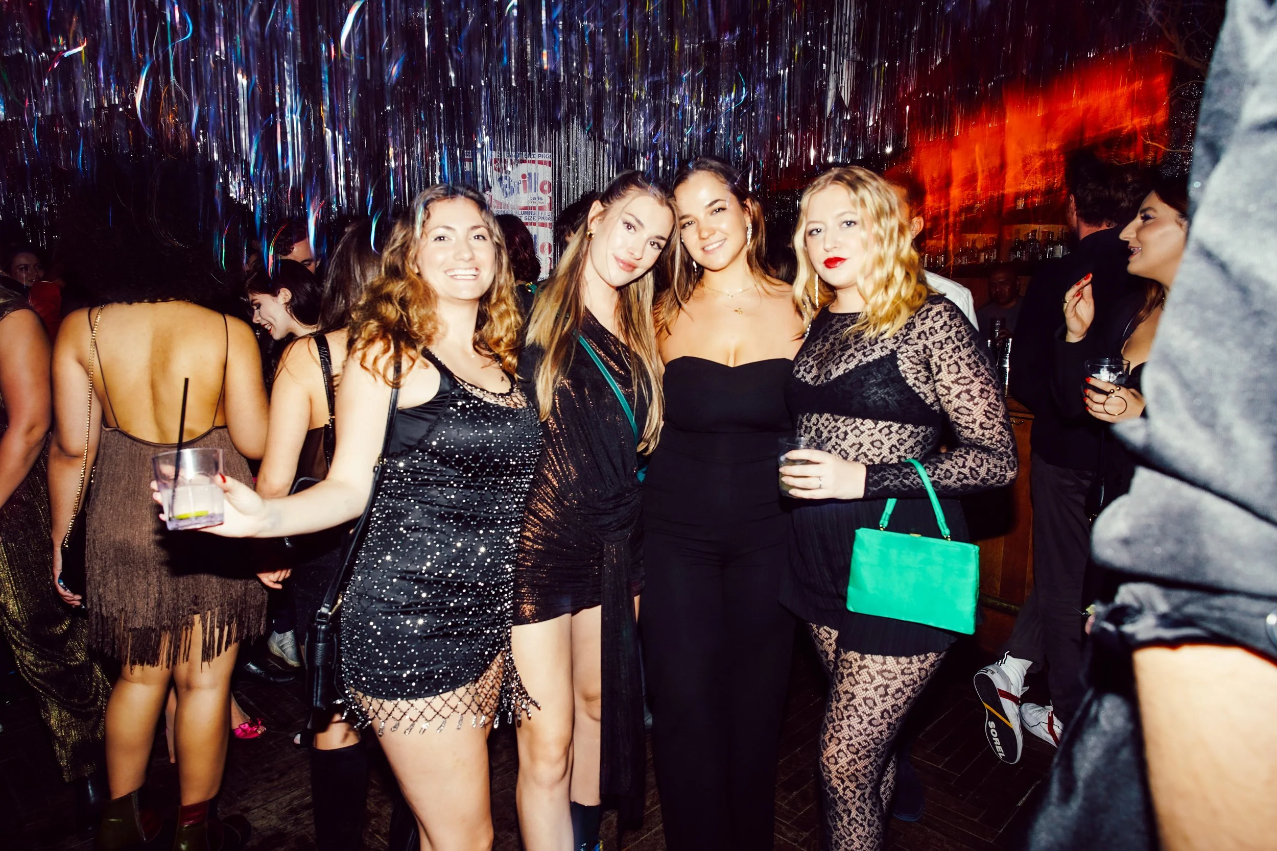 Group of women at a party, dressed in stylish black outfits, standing together and smiling for the camera.