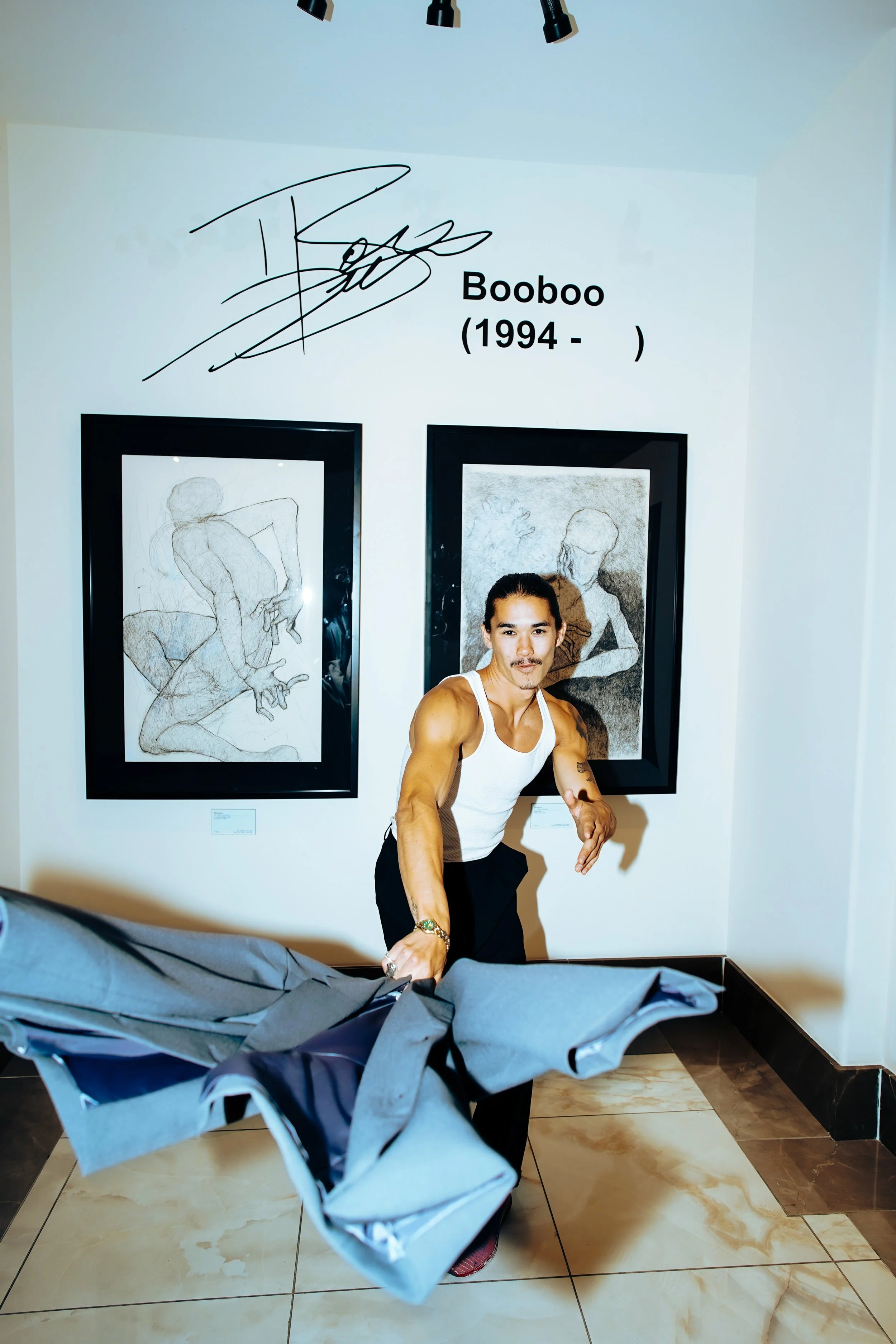 A man in a white tank top and black pants holding a gray jacket in an art gallery. Behind him are two framed sketches of nude women on a white wall, with a large signature and the name Booboo (1994-) written above.