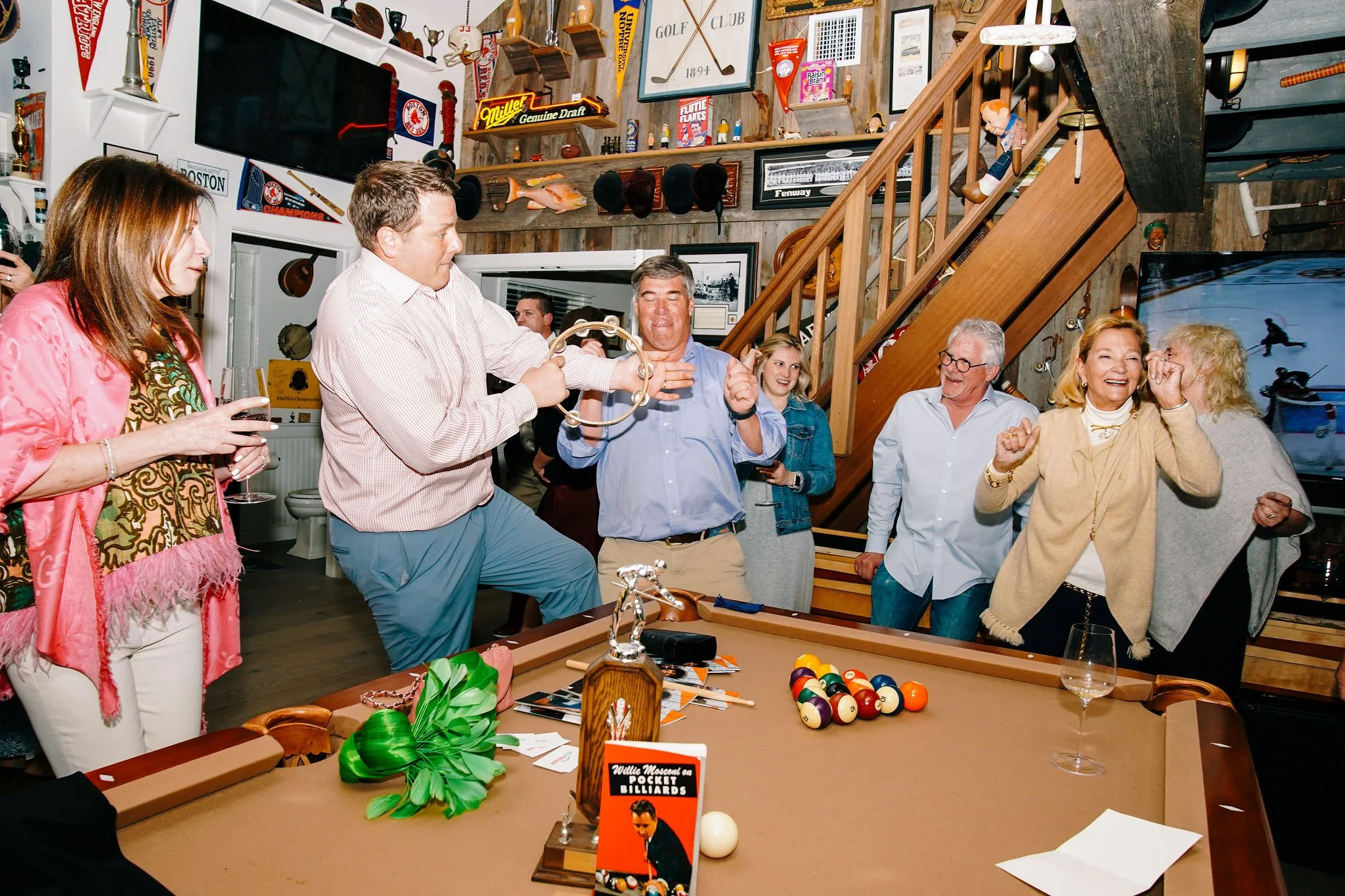 Group of people celebrating around a pool table in a wooden decor room with sports memorabilia, watching a man play a tambourine, while others cheer and laugh.
