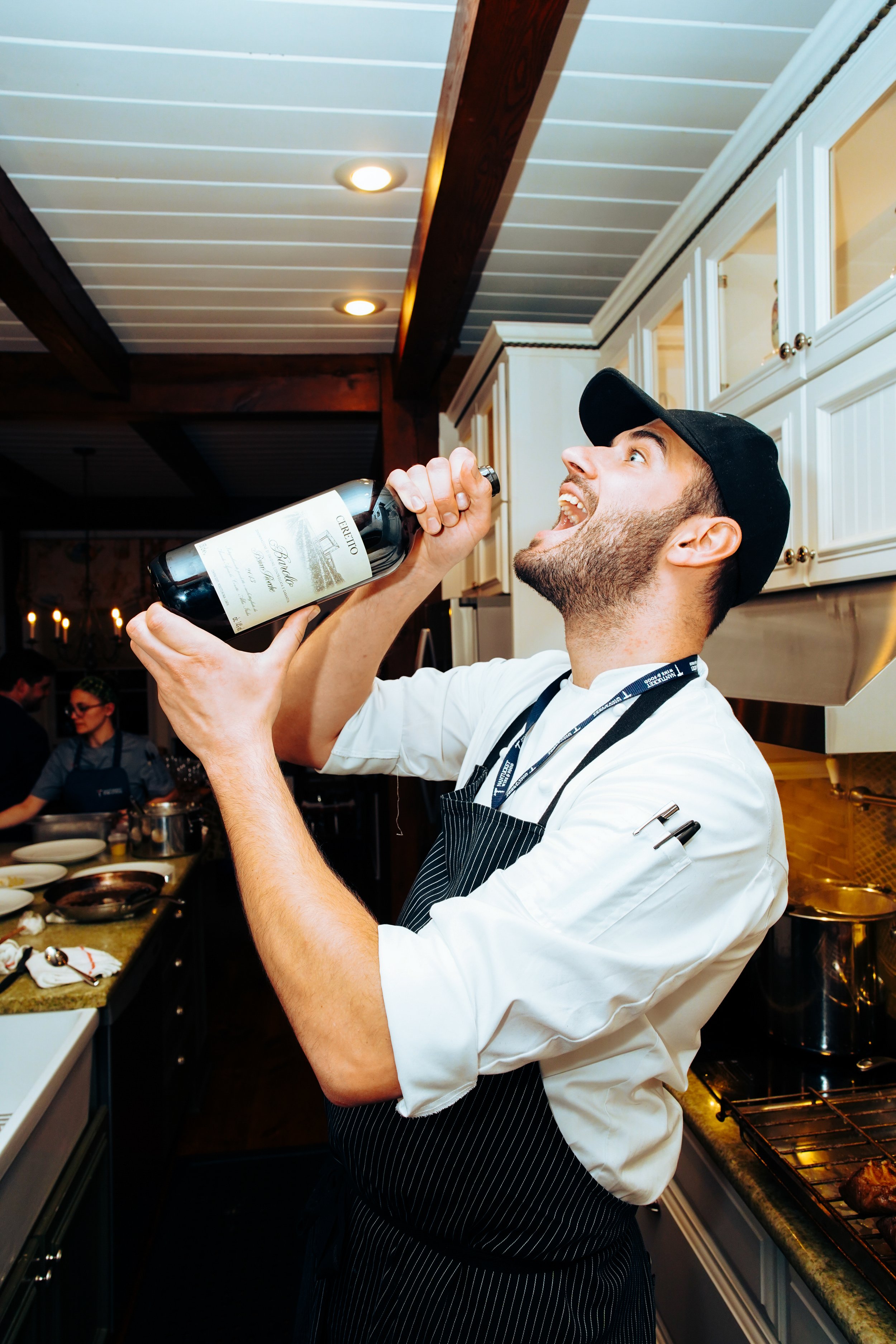A young man in a chef's uniform and apron drinking from a bottle of wine in a kitchen.