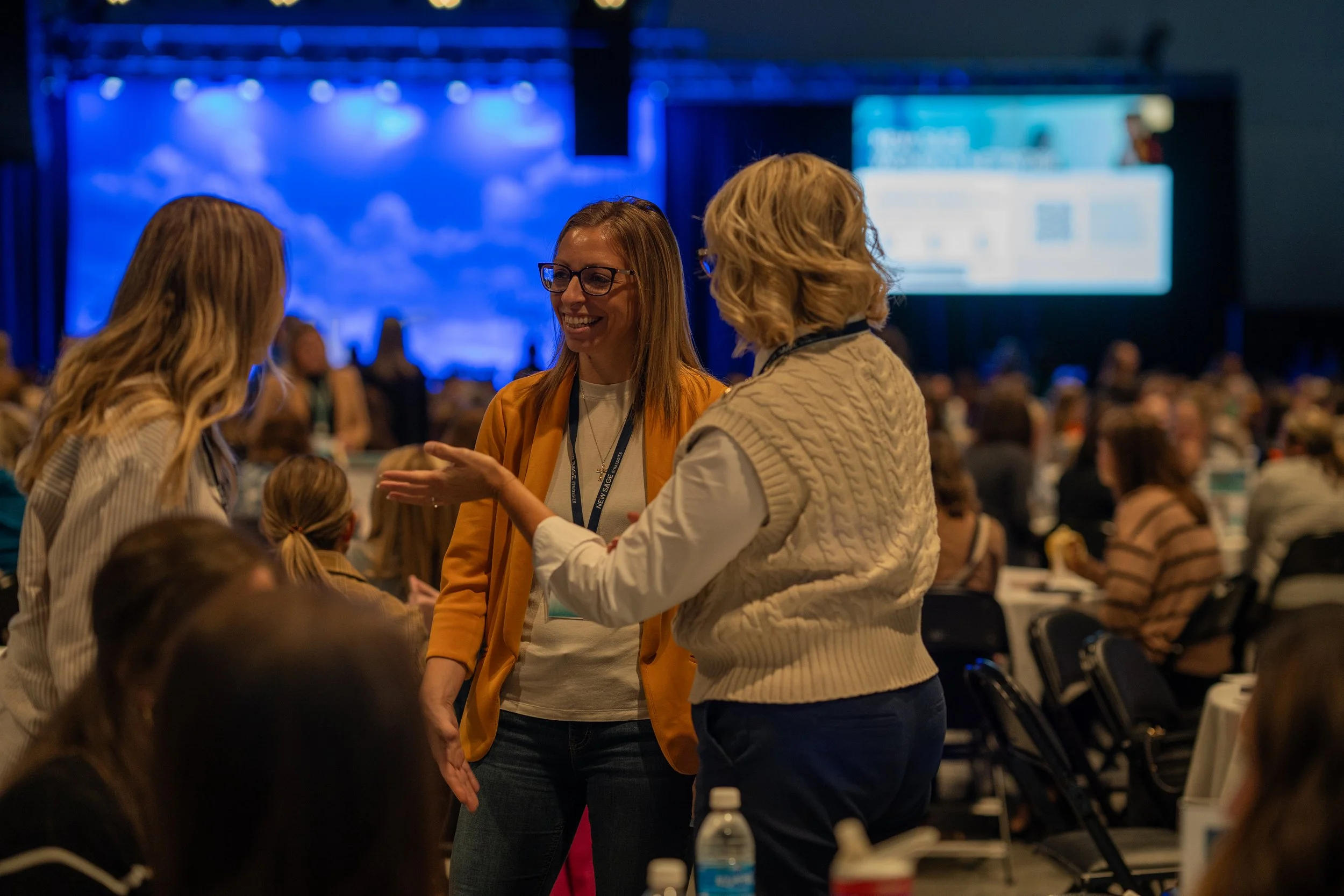 A group of people having a conversation in a conference setting with a stage in the background.