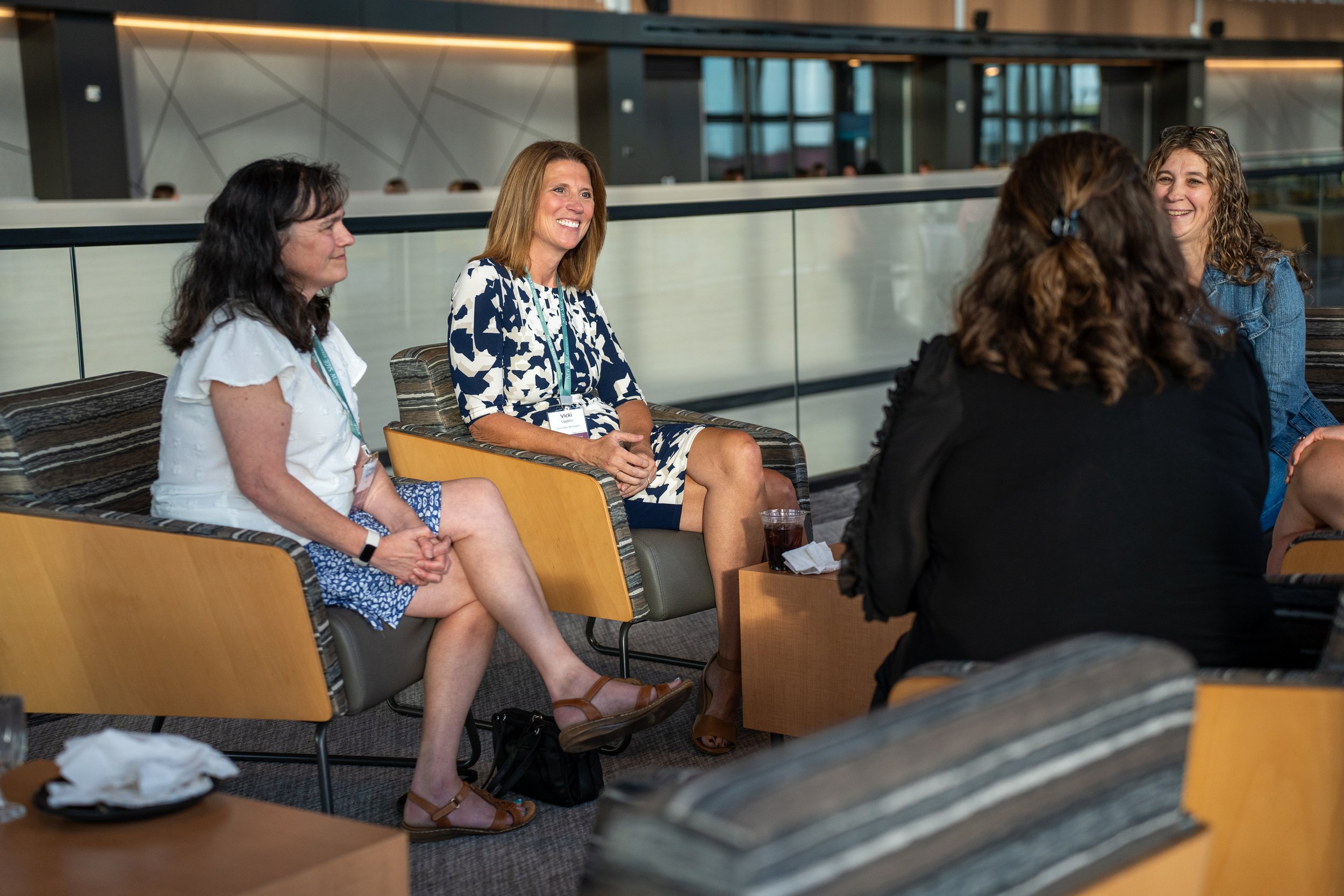 Four women sitting in a lobby or waiting area having a conversation, with modern furniture and a glass railing in the background.