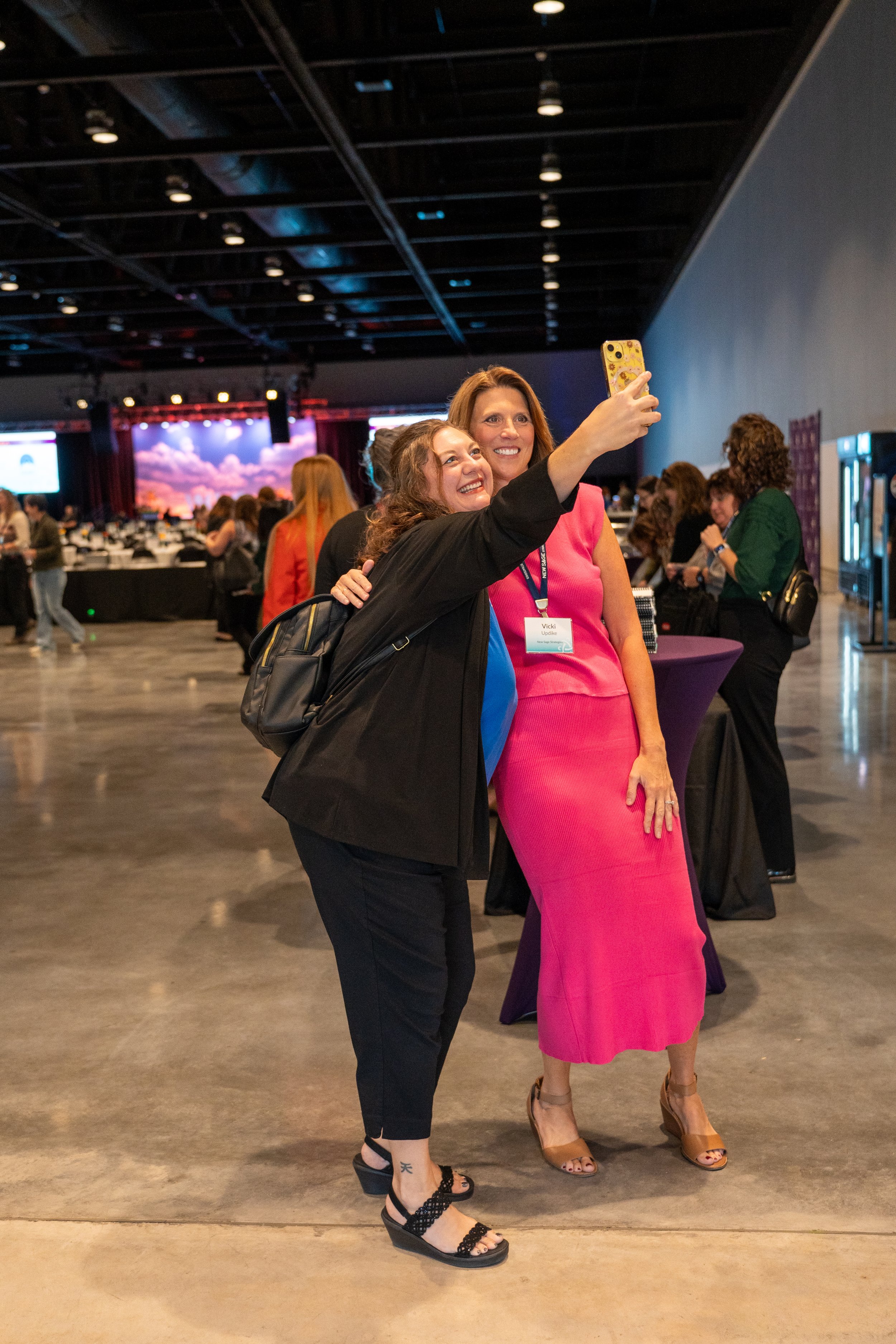 Two women taking a selfie at an indoor event, with one woman wearing a pink dress and the other in dark clothing. The background shows people at tables and event decorations.