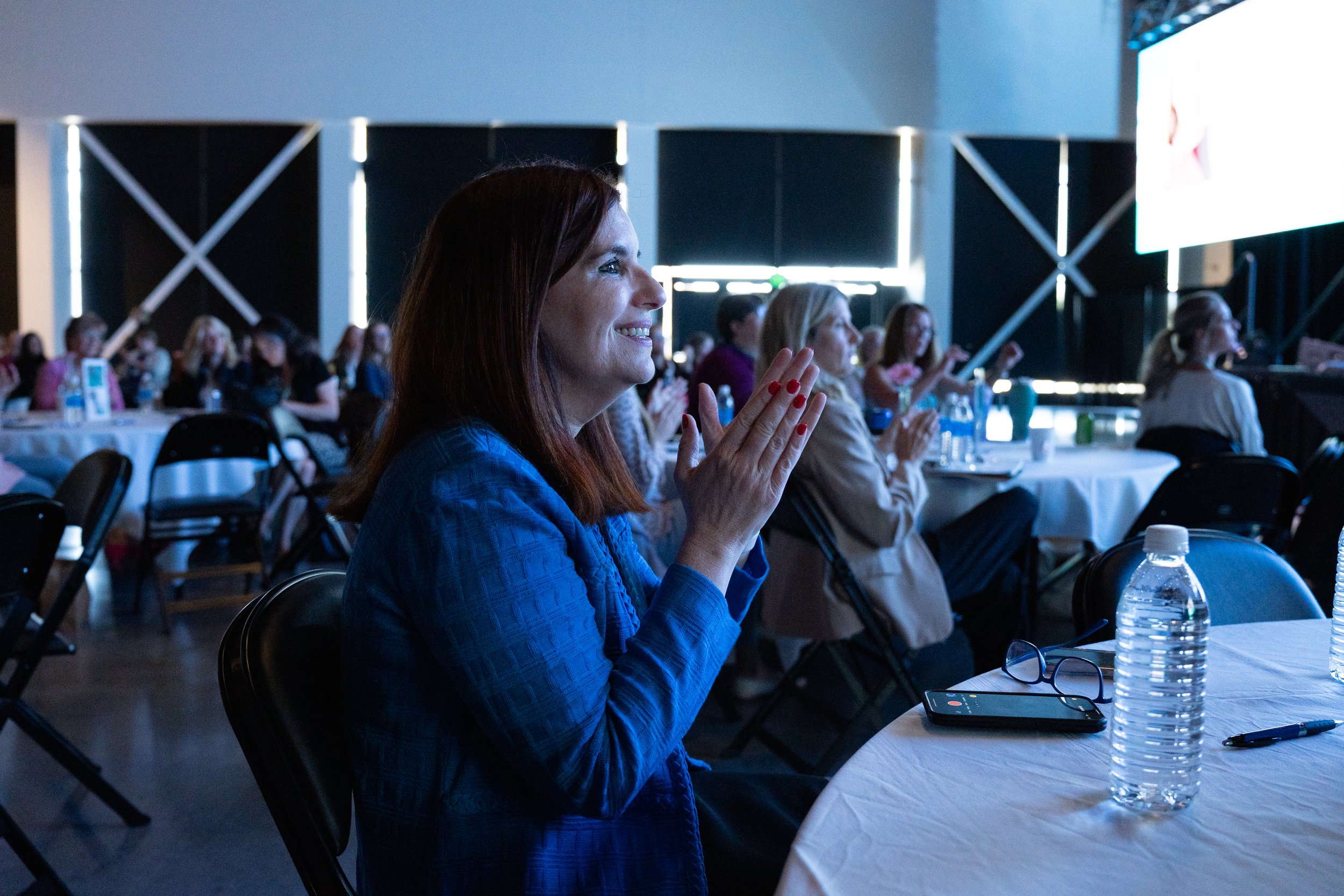 Audience clapping at a conference, woman in focus wearing a blue jacket, seated at tables with bottled water and conference materials.