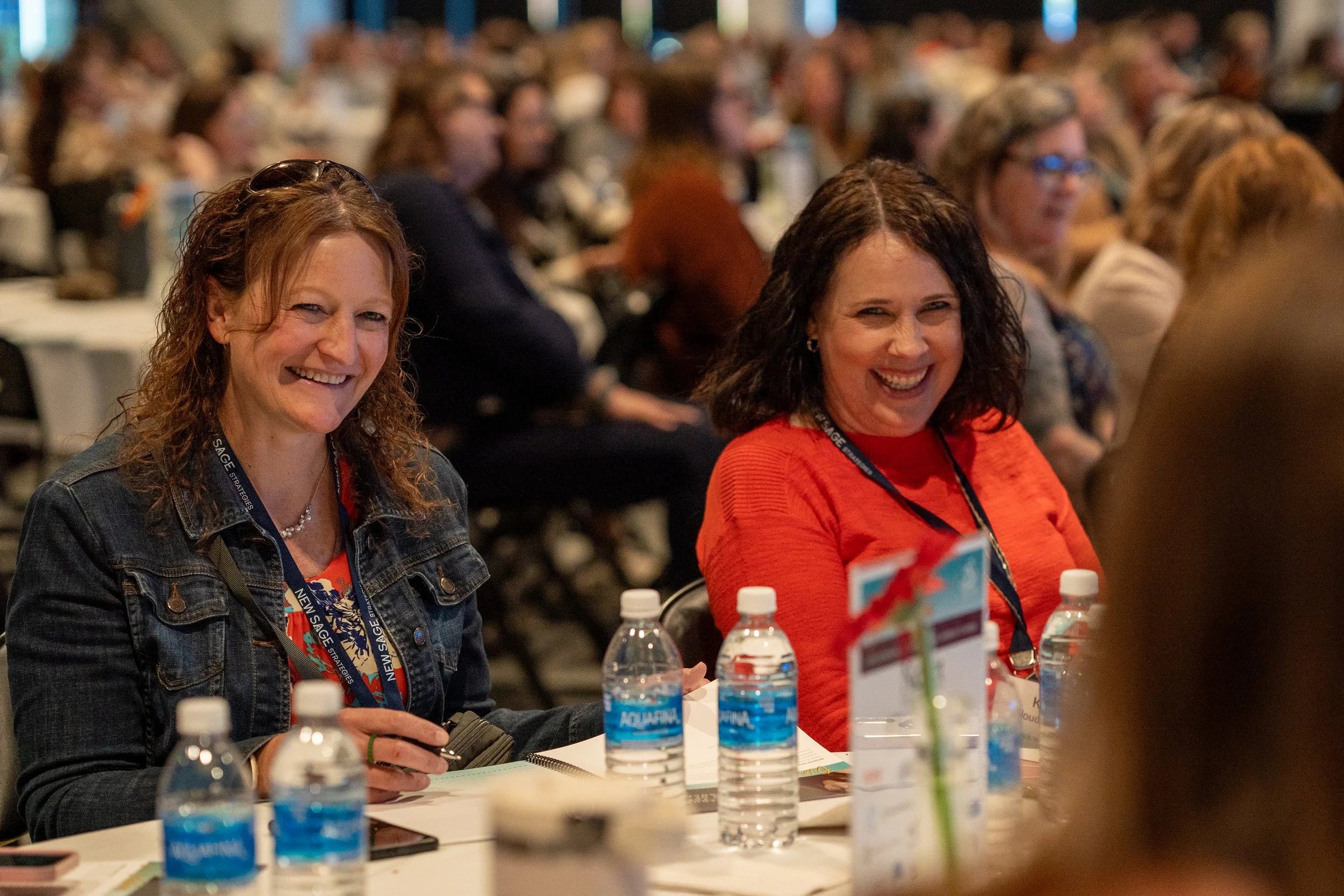 Two women smiling and sitting at a conference table with water bottles and papers, surrounded by a crowd of attendees.