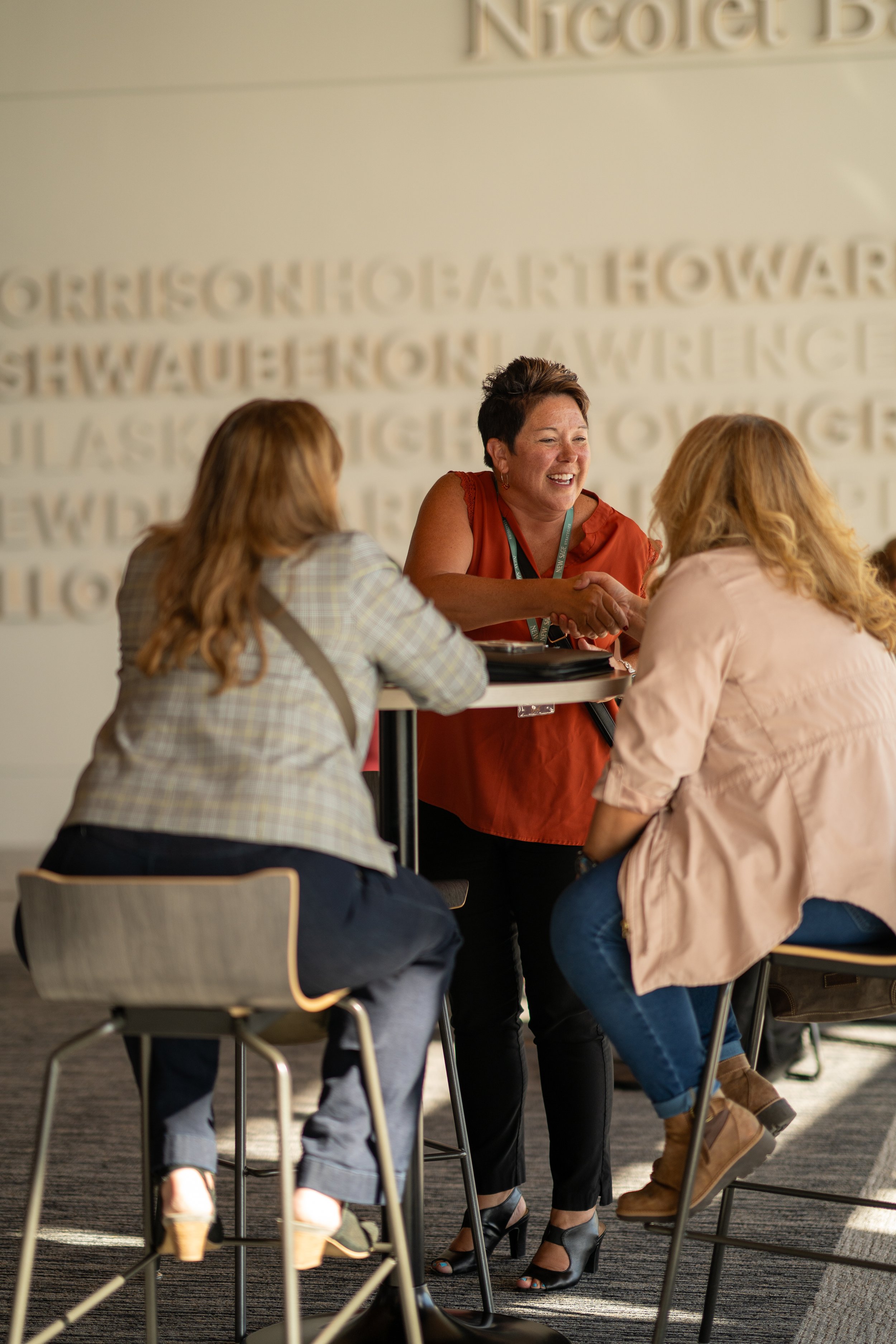 Three women seated around a high table having a conversation, with one woman smiling and engaging with the others.