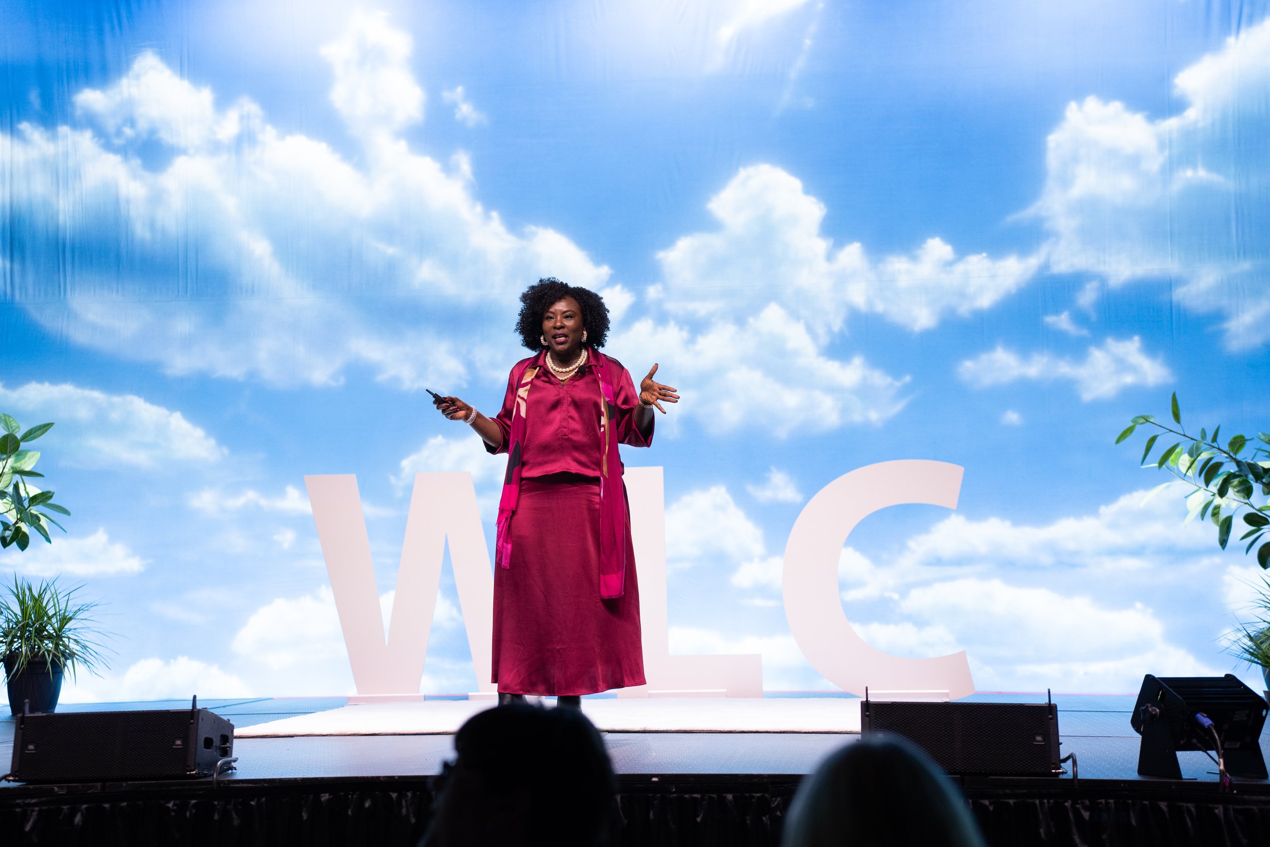 Person in pink outfit speaking on stage with a blue sky and clouds backdrop, large letters "WLC" in the background.