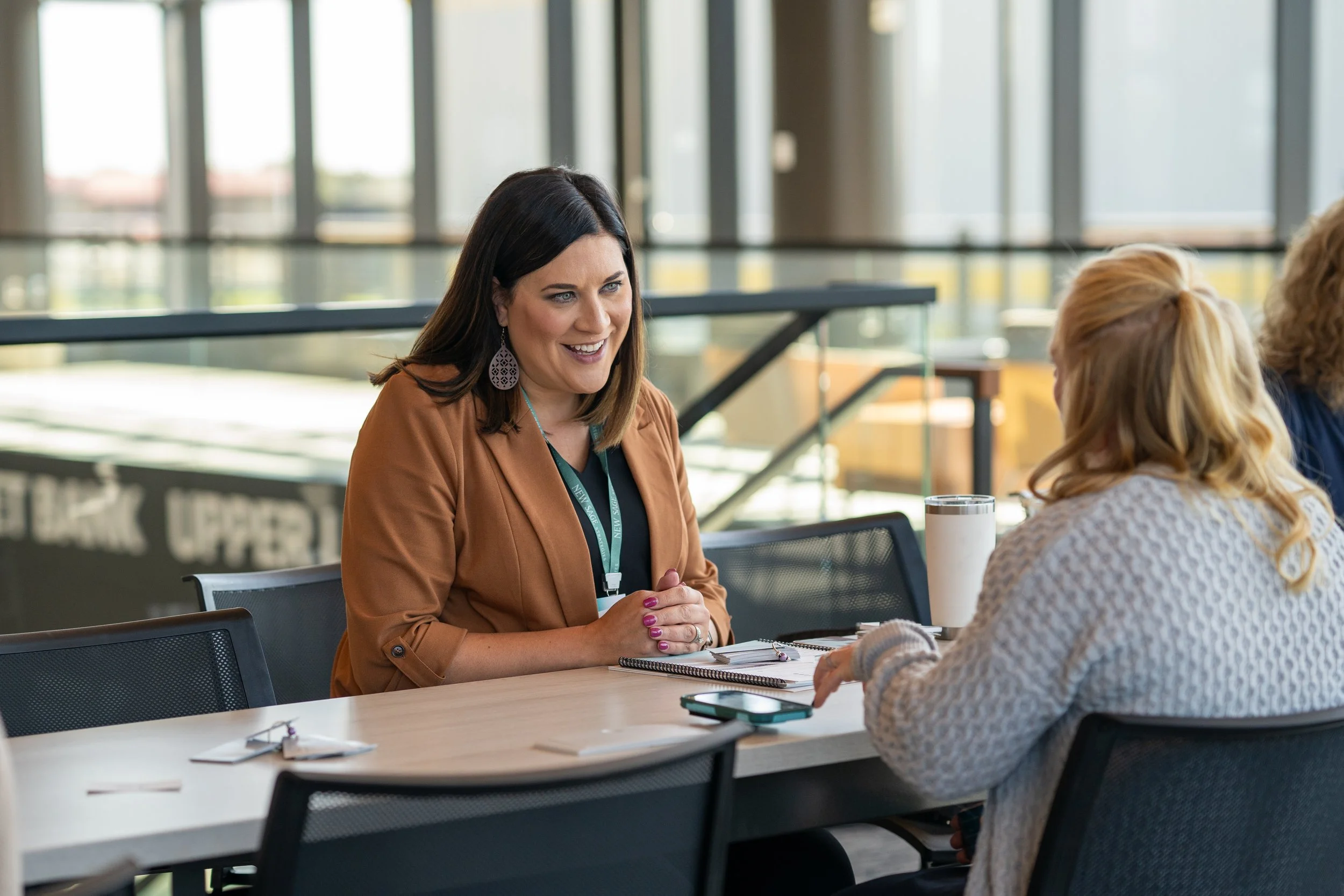 Two women sitting at a table, engaged in conversation in a modern office setting.