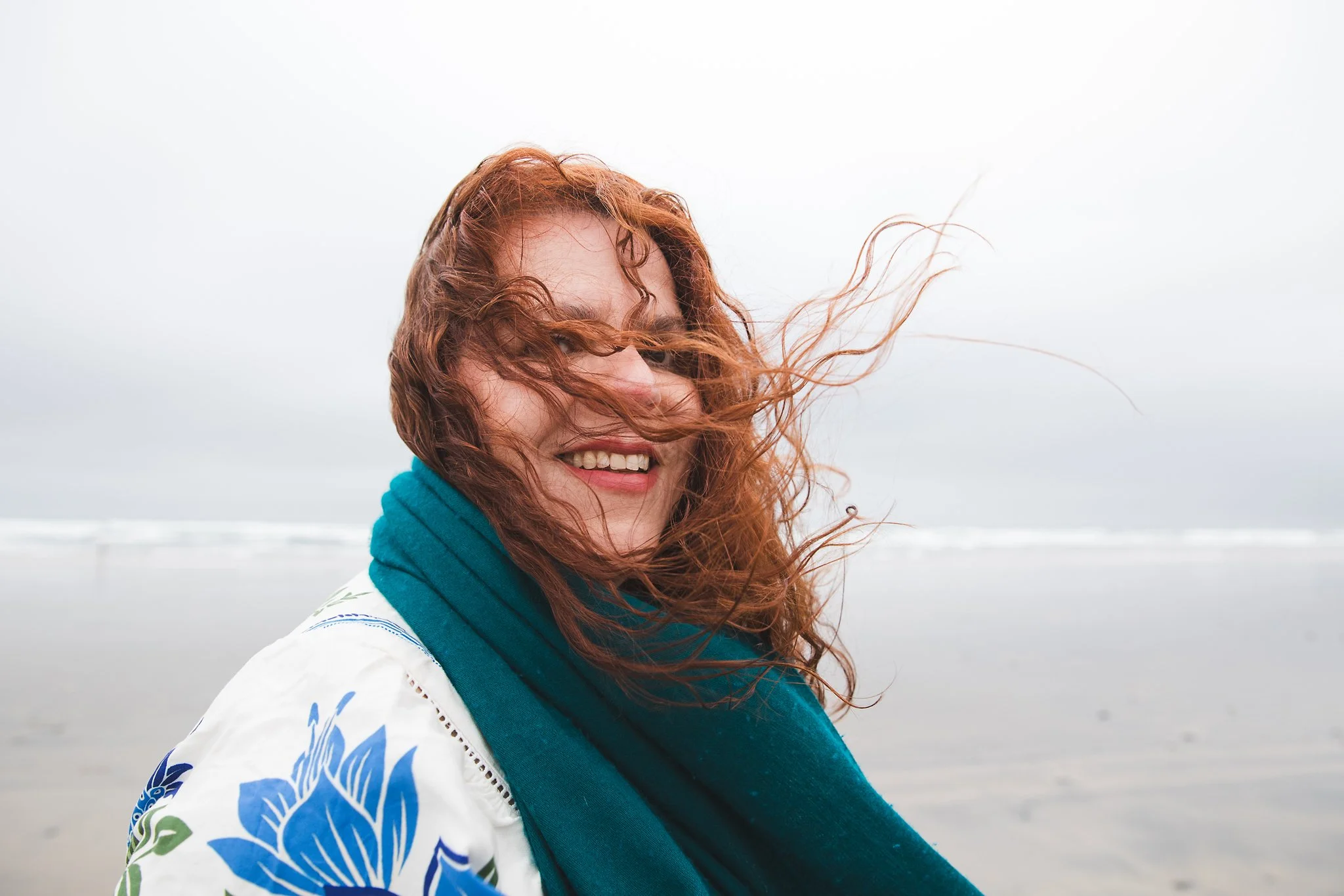 A woman with red hair and a blue scarf smiling on a windy beach during overcast weather.