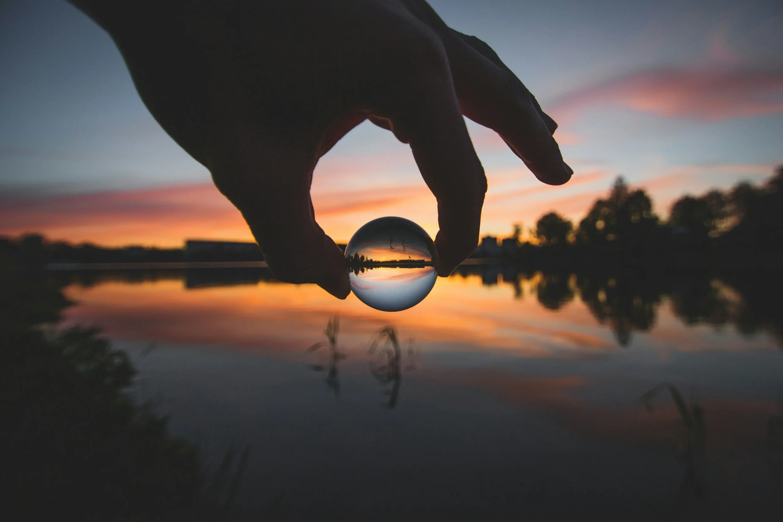 Person holding a glass ball reflecting a sunset over a lake with trees and colorful sky.