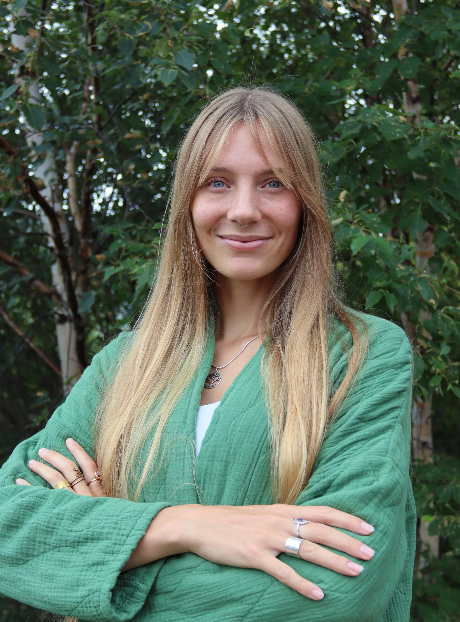 A woman with long blonde hair and blue eyes smiling outdoors, crossing her arms, in front of a background of green leafy trees.