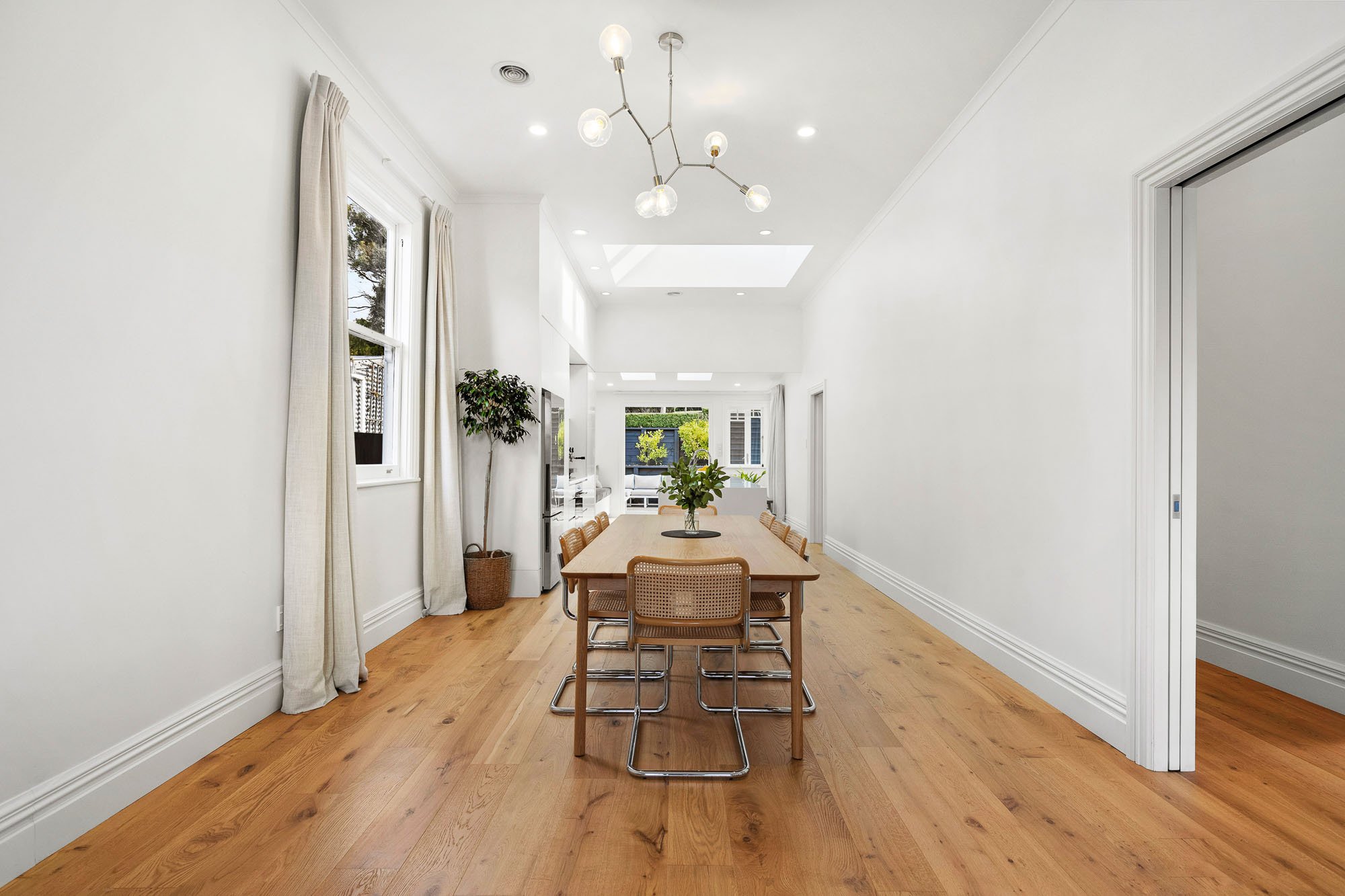 Modern dining room in Wellington, NZ, with wooden table and chairs, hardwood floor, large windows with beige curtains, potted plant, contemporary ceiling light, and bright, open layout. Designed by Kate Harford from Hark Architects