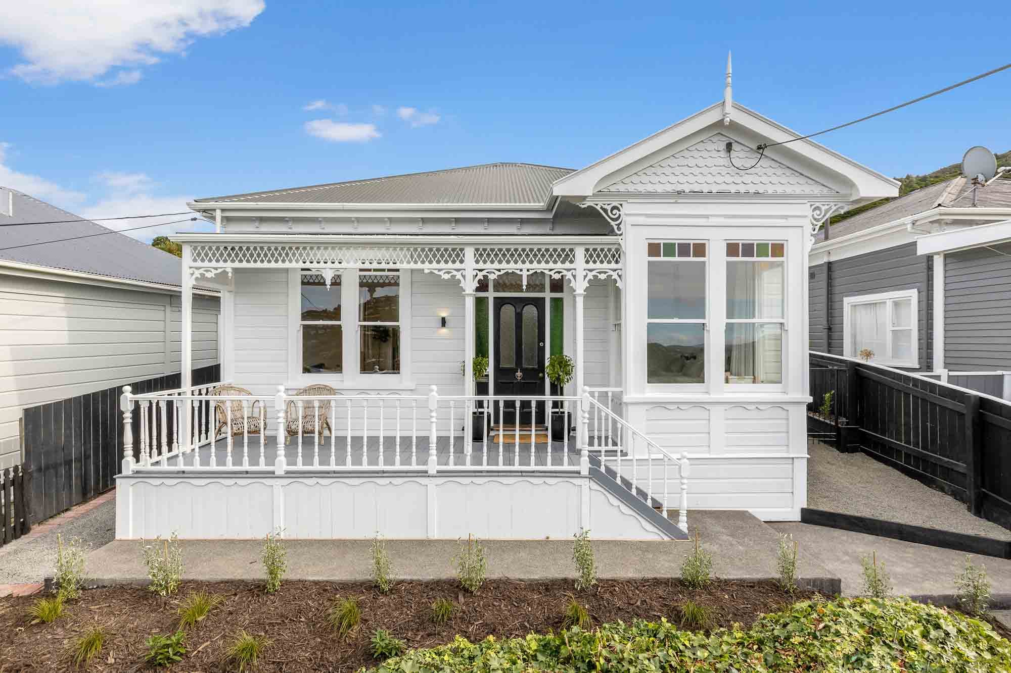 Victorian-style white villa with ornate trim, a covered porch, wicker chairs, and landscaped yard. The interior of this home was renovated and remodelled by Hark Architects, a Wellington architectural firm and registered architect.