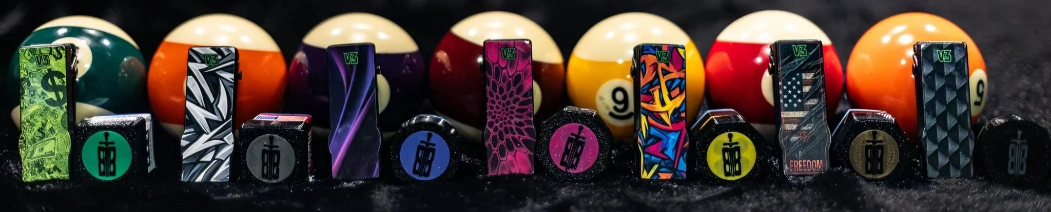 Collection of V3 magnetic chalk holders with various designs, lined up in a row on a black background, with pool balls in the background.