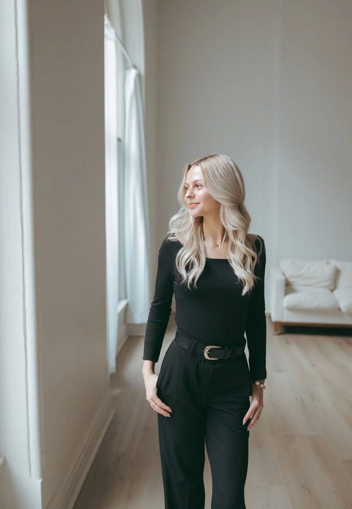 A woman with long blonde hair, wearing a black long-sleeve top and black pants with a belt, standing in a bright room near a window with white curtains, looking to her left and smiling.