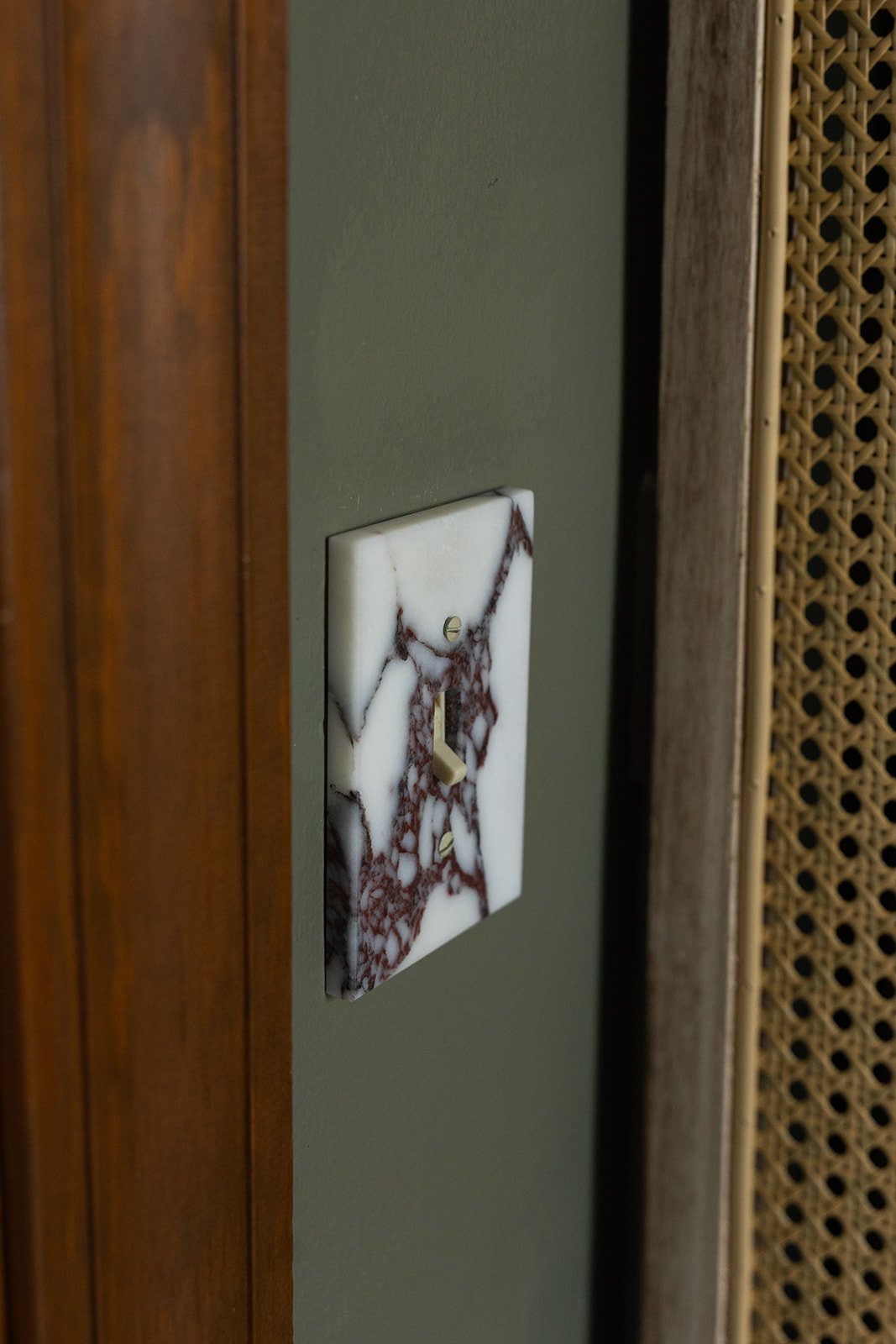 A light switch with a marble design cover plate on a green wall, near a wooden door and a woven rattan panel.