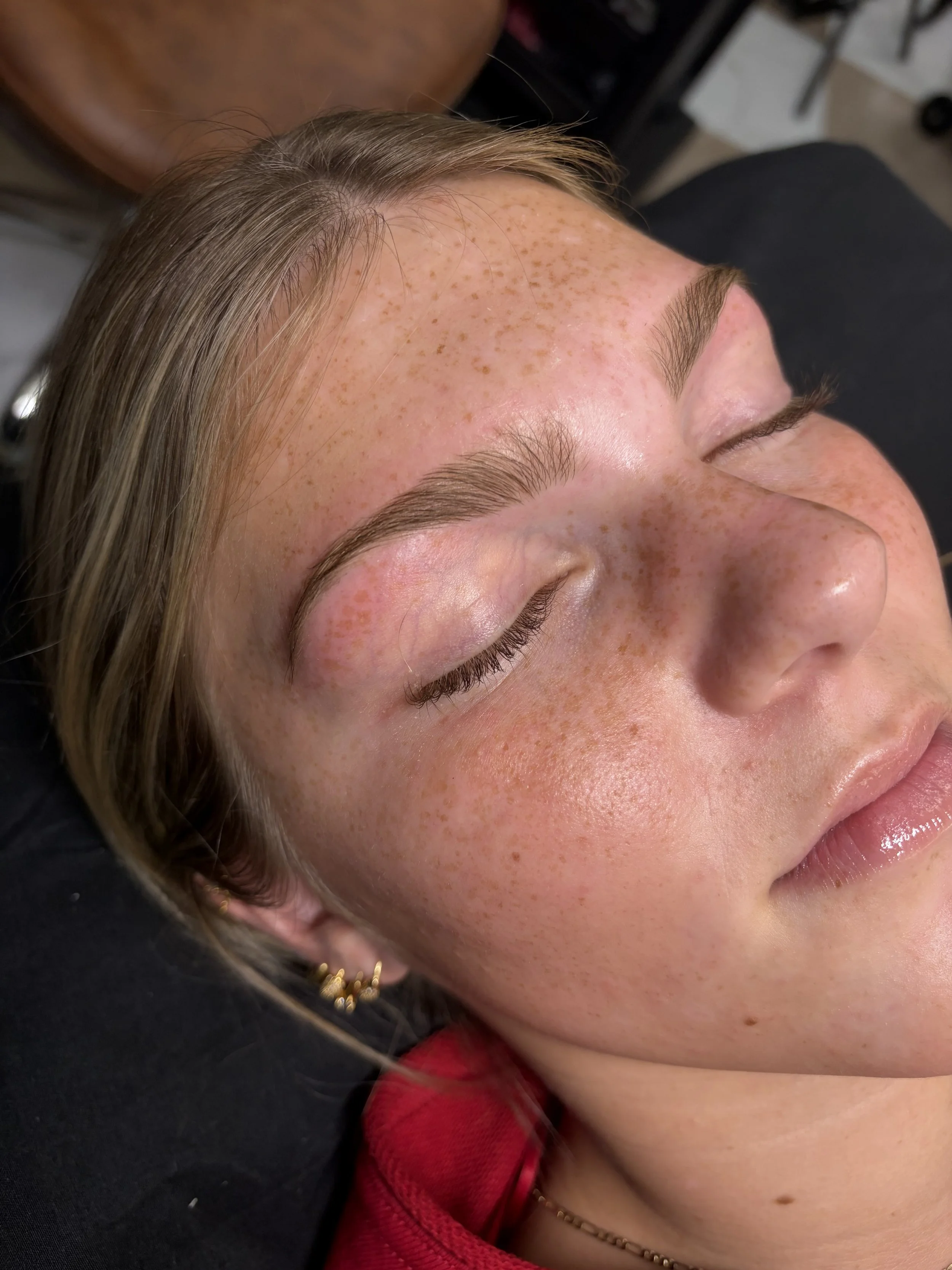 Close-up of a woman with closed eyes, showing her face with freckles, smooth skin, and well-groomed eyebrows. She is lying down, possibly at a beauty or skincare treatment.