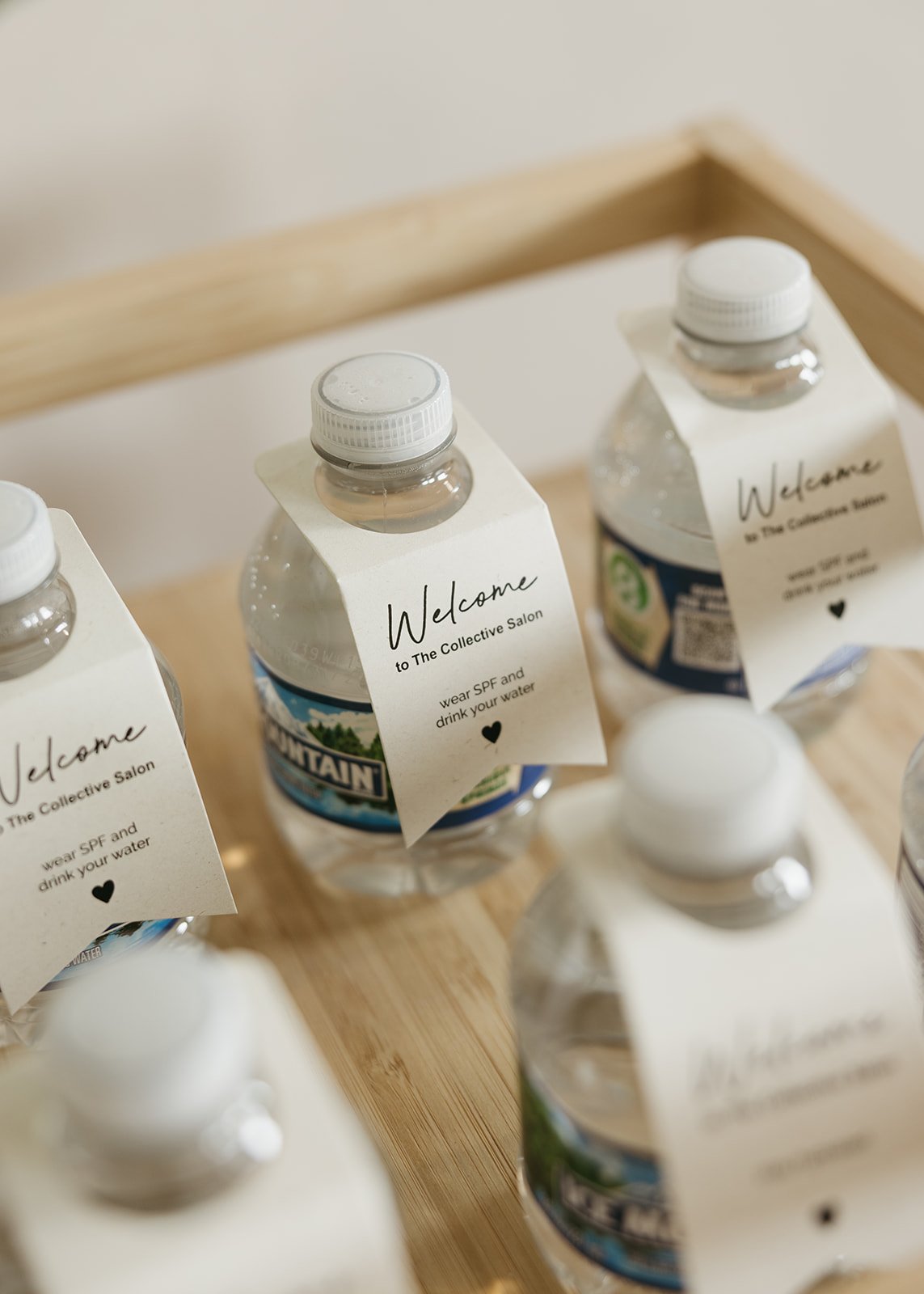 Collection of bottled water with tags that say "Welcome to The Collective Salon" and "wear SPF and drink your water" on a wooden tray.