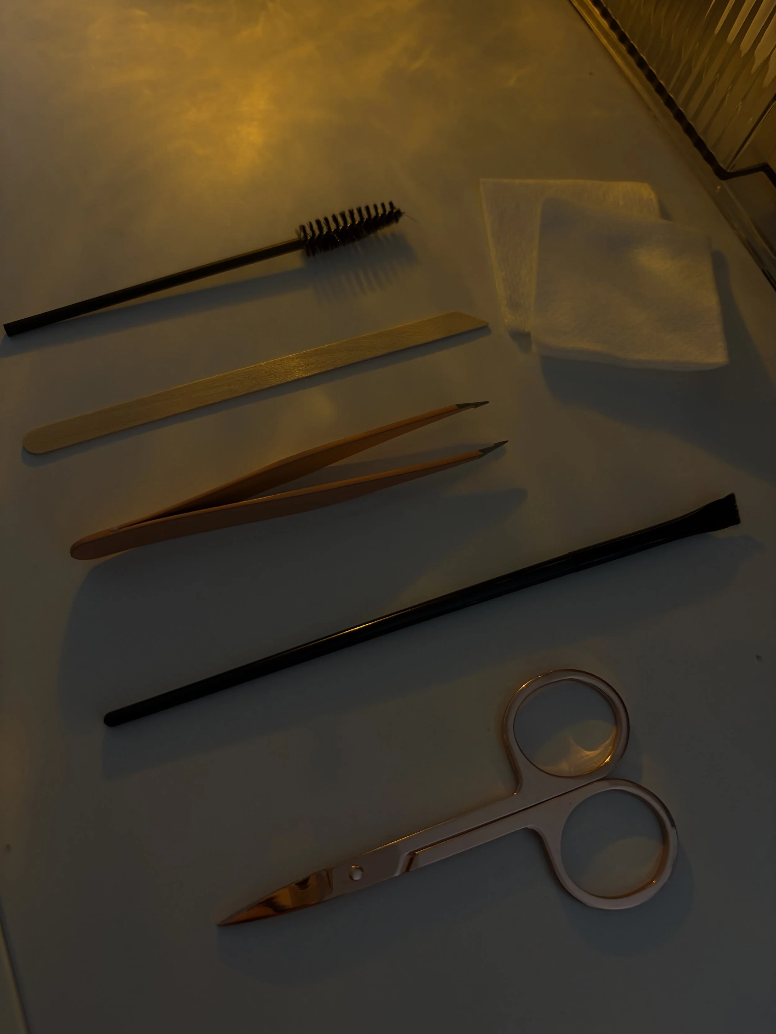 A collection of beauty tools including a spoolie brush, cotton pads, wooden swabs, tweezers, and scissors placed on a white surface.