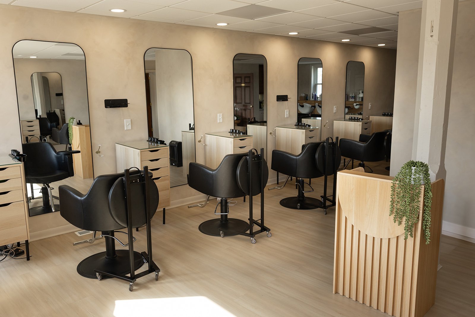 Empty hair salon with four styling stations, each with a mirror, black styling chair, and wooden drawers, arranged in a row on a light wood floor.