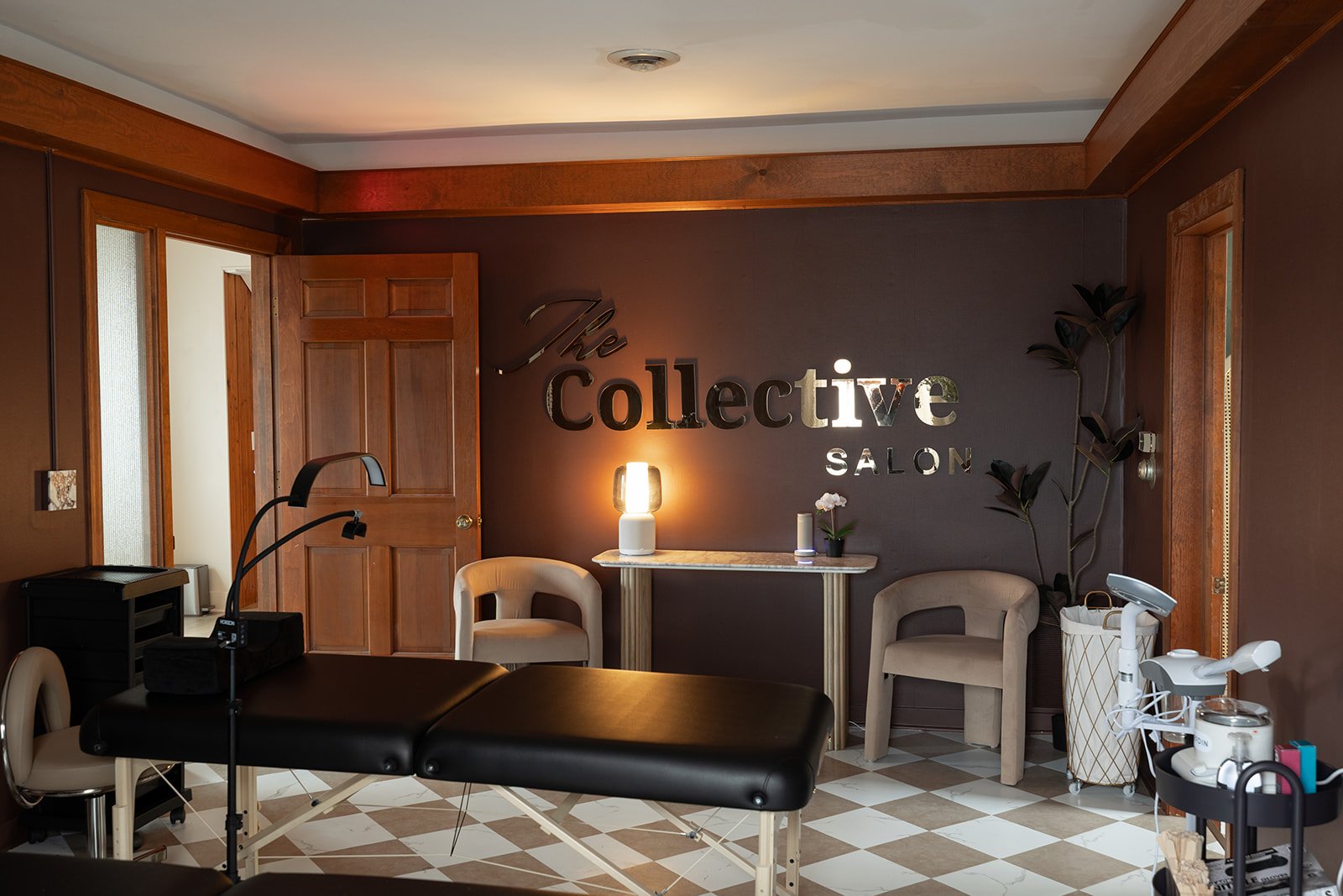 Interior of a salon with a black massage table, beige chairs, a marble-topped table with a lamp, small plant, and decorative wall sign reading 'The Collective SALON' on a brown wall.