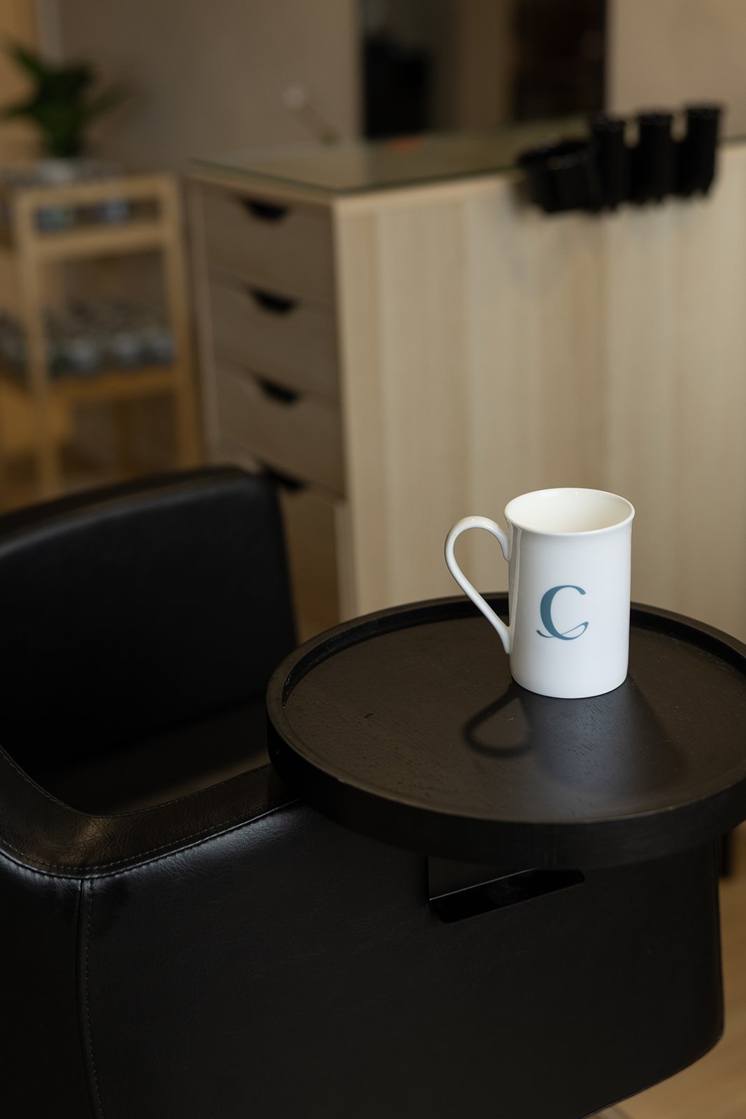 White coffee mug with a C on it placed on a black circular tray, on top of a black leather chair.