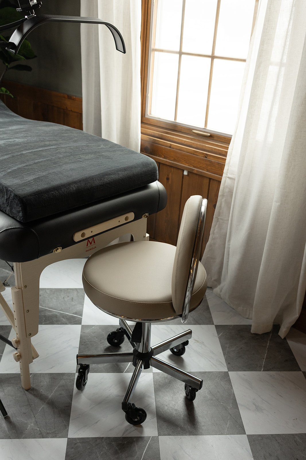 View of a black massage table and a beige rolling chair by a window with white curtains and wooden panels.
