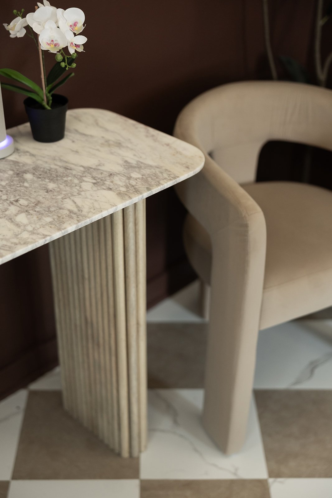 A marble table with vertical, ribbed beige_column support, topped with a potted orchid, and a cream-colored, upholstered armchair with rounded armrests next to it.