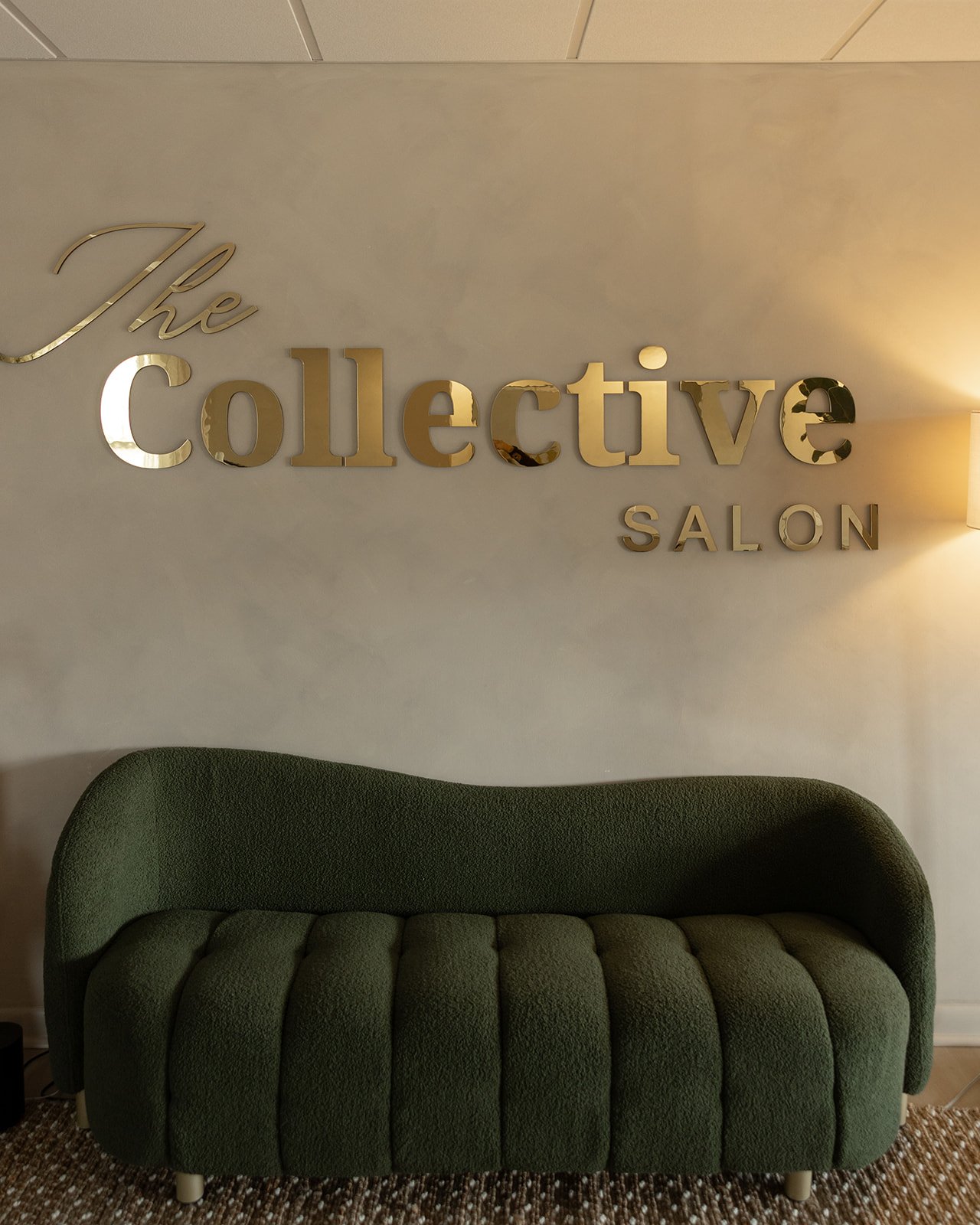 Green upholstered sofa in front of a wall with gold letters reading 'The Collective Salon' and a wall-mounted light on the right.