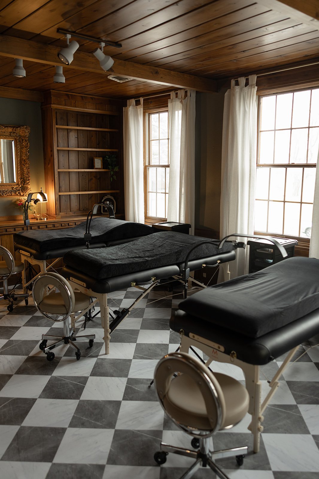 A room with two massage tables, black linens, and adjustable lamps. The room has wooden walls, large windows with white curtains, and a checkered black and white floor.