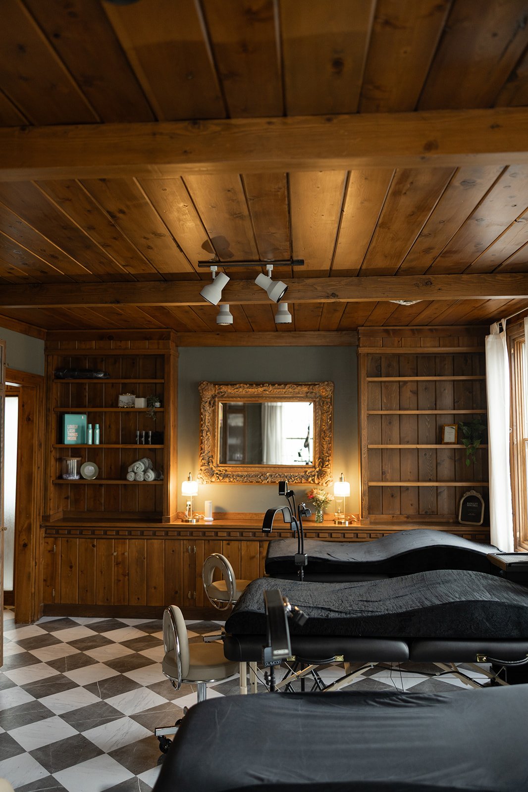 A cozy room with wooden walls and ceiling, a large mirror, two mirrored lamps, massage tables, and a checkered tile floor.