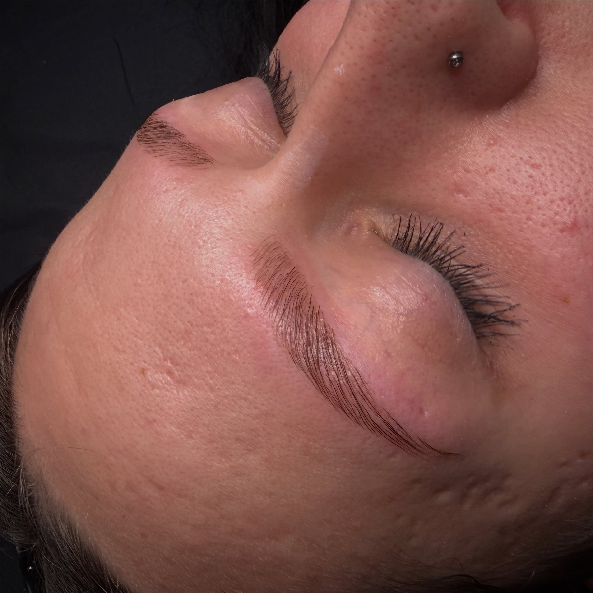 Close-up of a woman's face showcasing smooth skin, arched eyebrows, long eyelashes, and a nose with a small piercing.