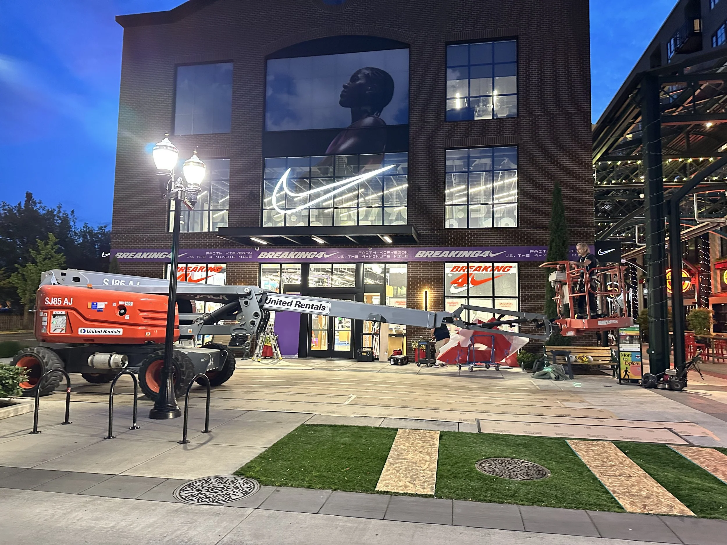 Construction workers setting up equipment outside a Nike store at dusk. The Nike logo is illuminated on the building's large windows, and a cherry picker lift is being used for work. The scene includes a lamppost, a sidewalk, and some greenery.