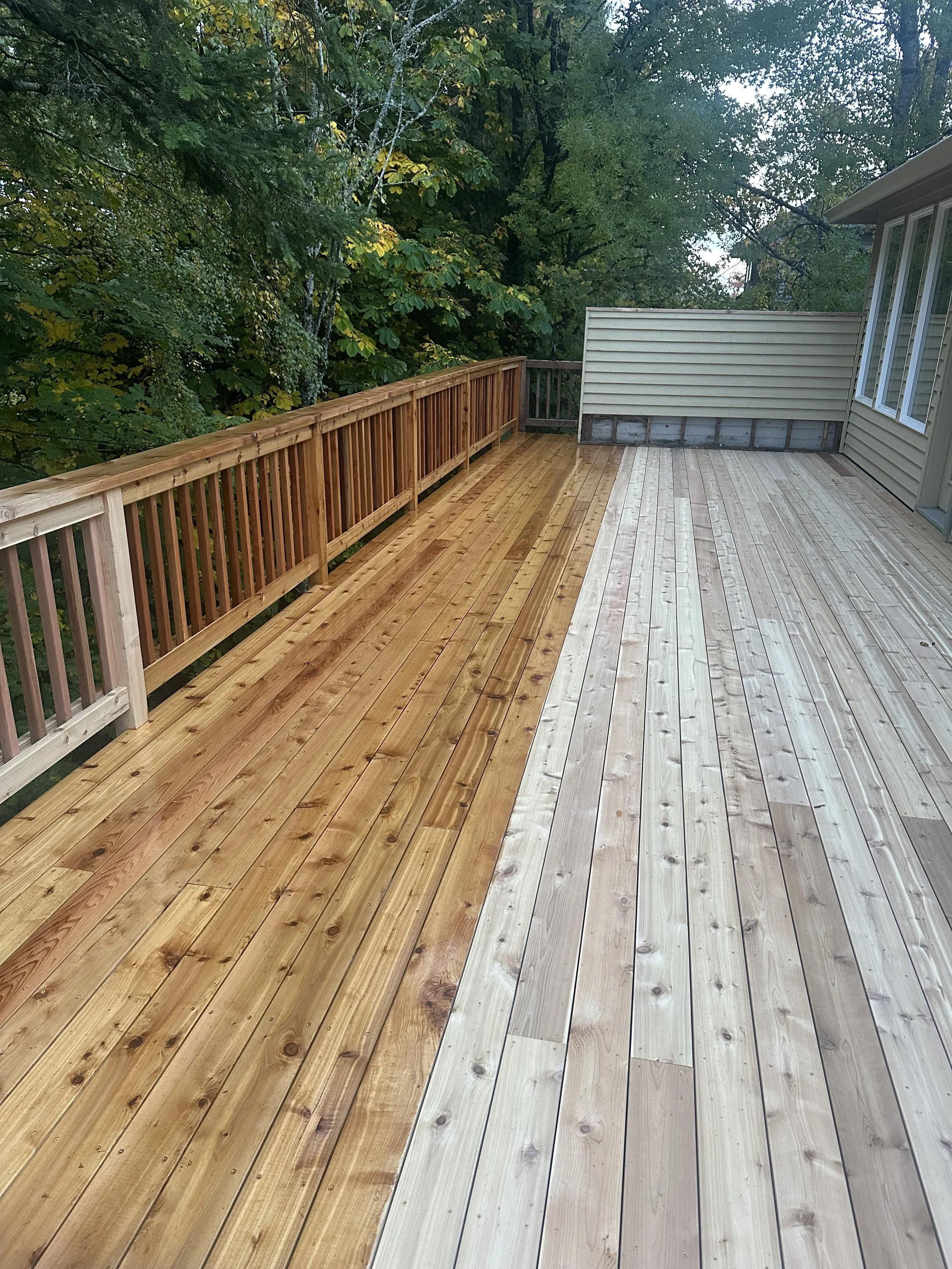 A newly built wooden deck with different staining on each side, bordered by a wooden railing, surrounded by trees with green leaves.