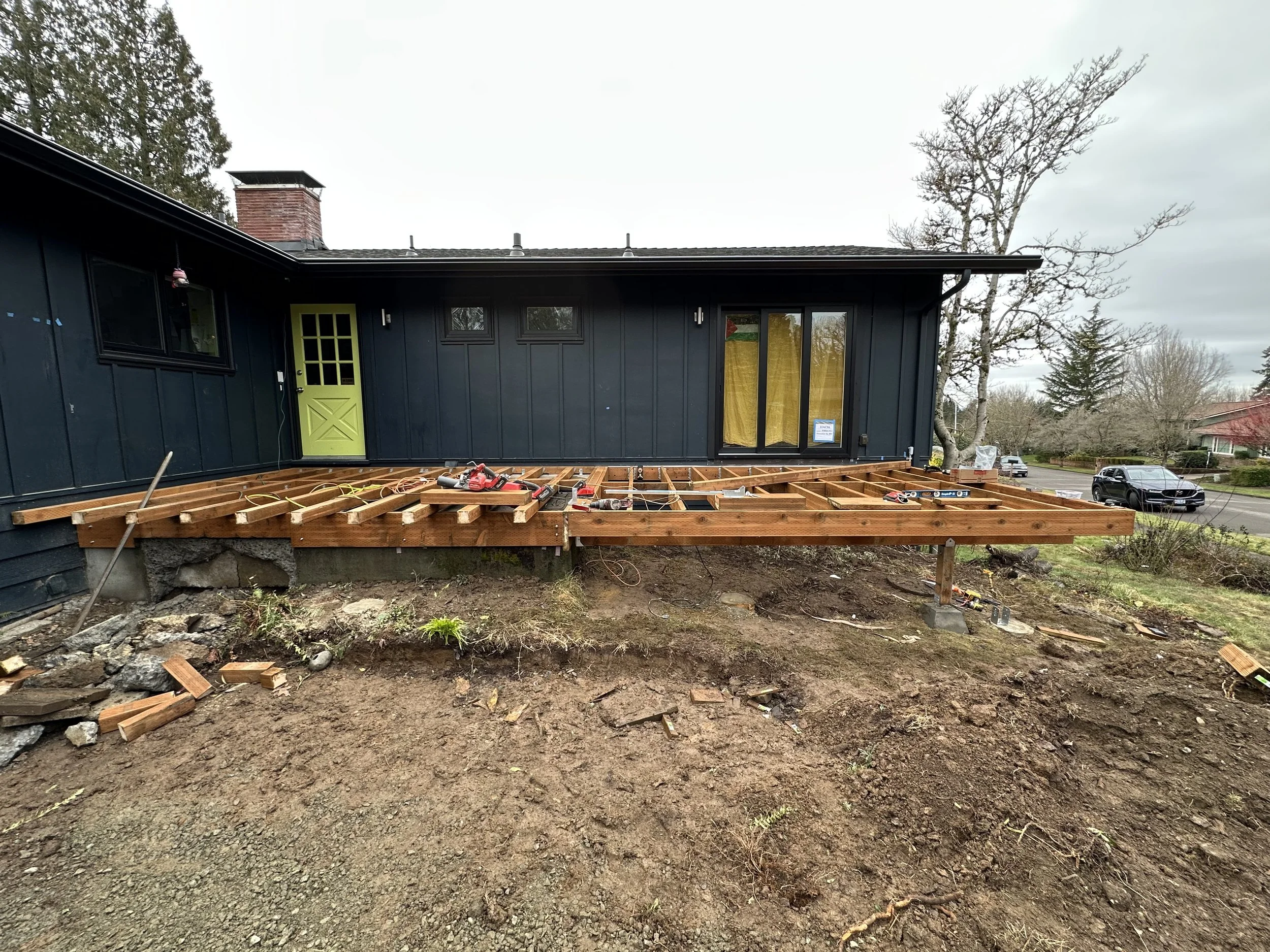 Construction site for a wooden deck attached to a house with black exterior, green door, and sliding glass door, showing the framing in progress.