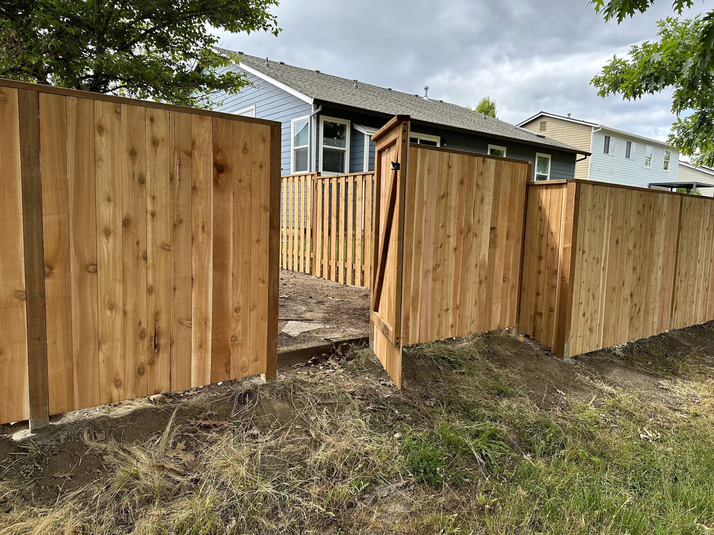 A wooden fence with an open gate in a backyard, with a blue house and trees in the background, under a cloudy sky.