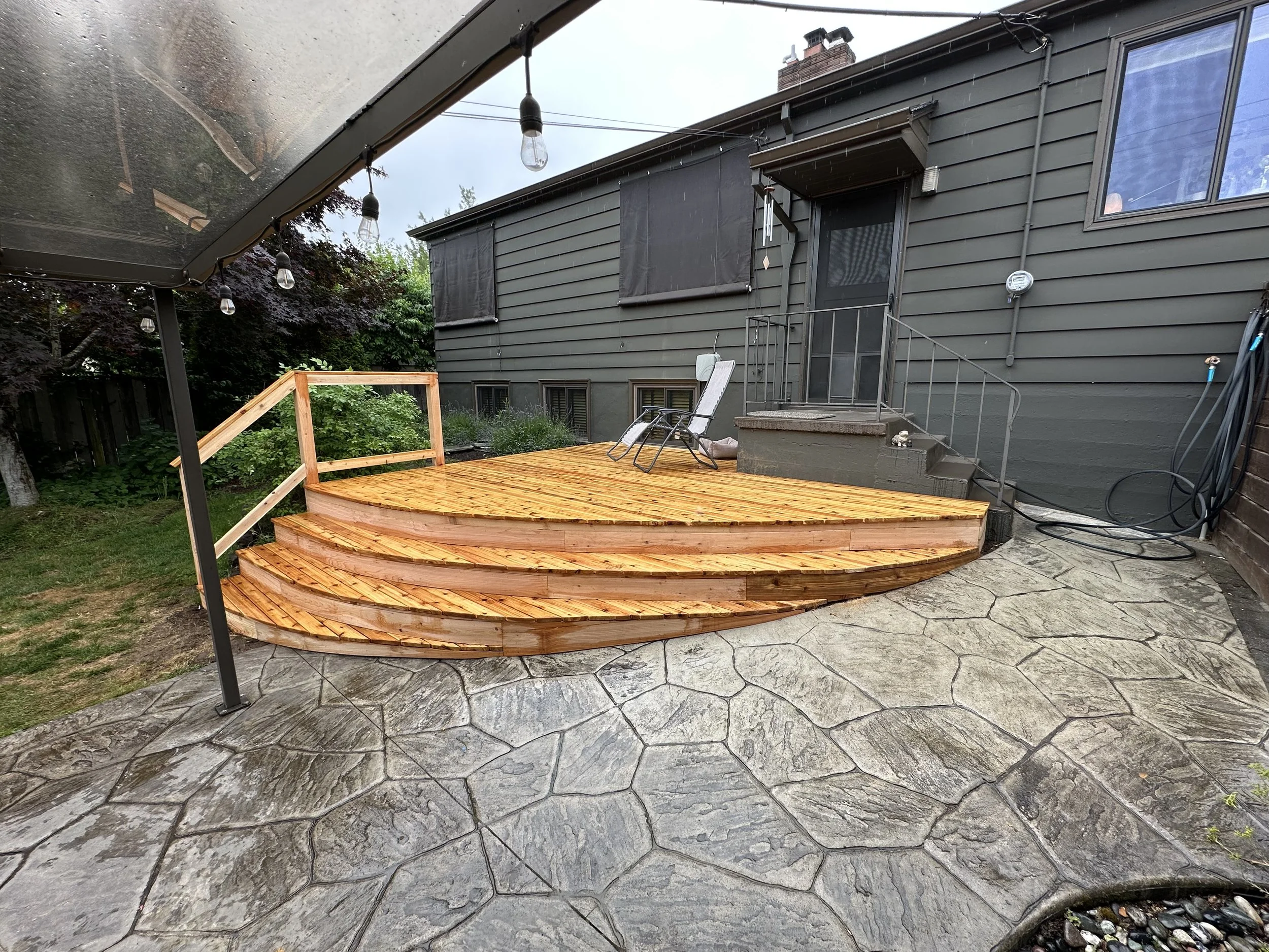 Wooden deck with four steps and a black metal railing next to a gray house with a black door and windows. The deck has a lounge chair and a pair of shoes. A string of hanging light bulbs is visible under a patio cover. The ground is concrete with a stone pattern, and there's a garden bed with plants on the left side.
