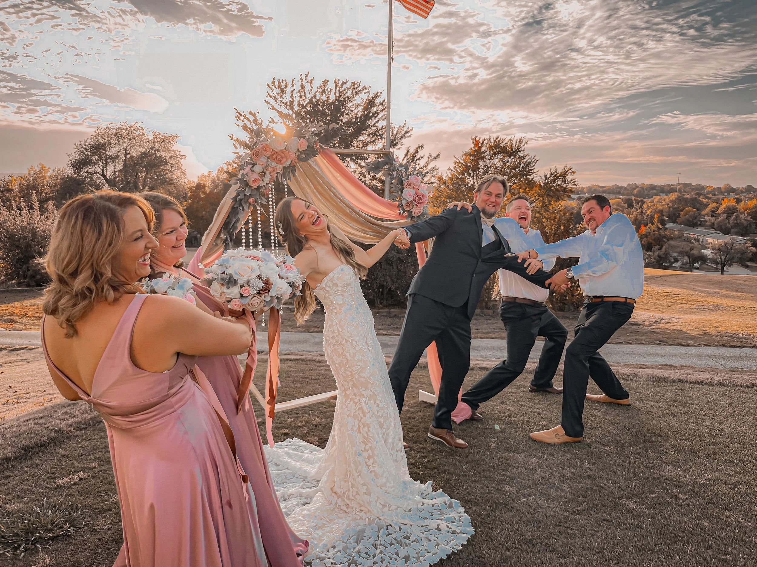 Group of people holding hands and dancing in a circle outdoors during a wedding celebration at sunset, with decorated arch and trees in the background.