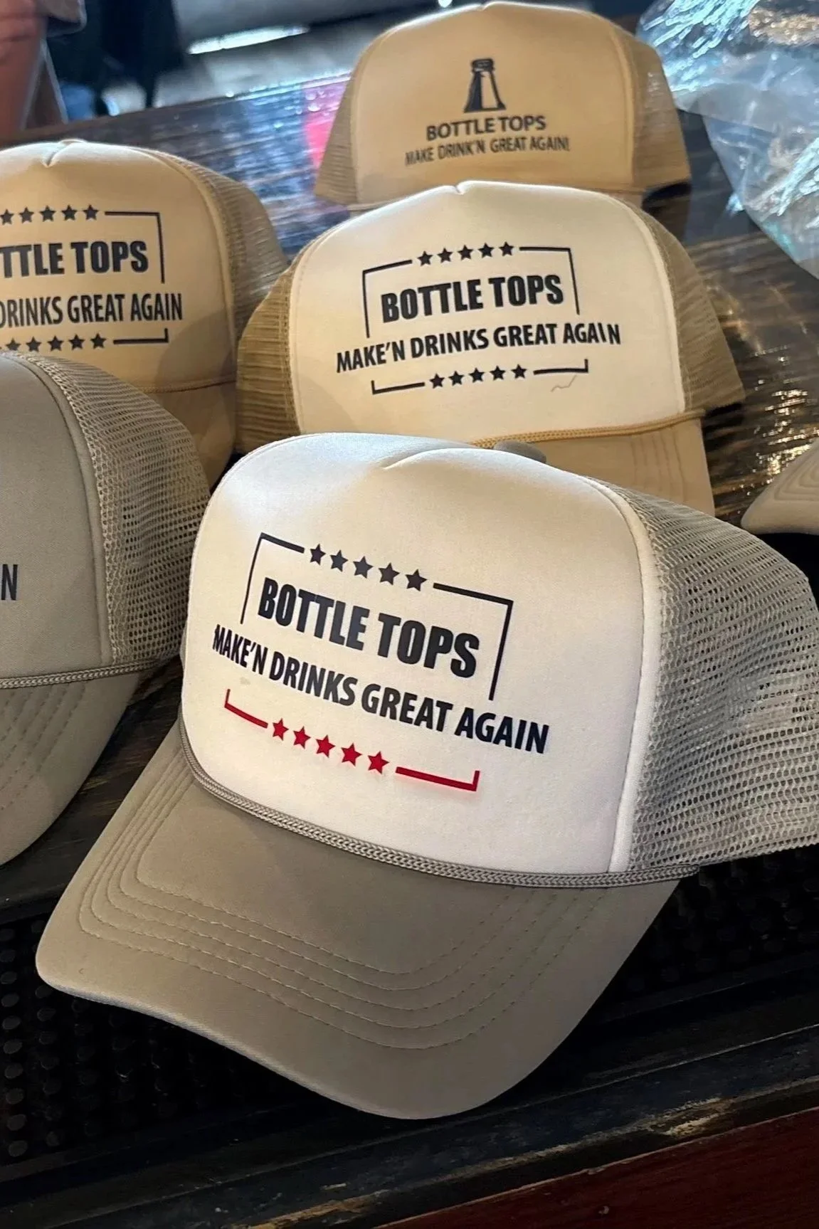 Several white baseball caps with black and red text reading "Bottle Tops Make 'n Drinks Great Again" on a table.