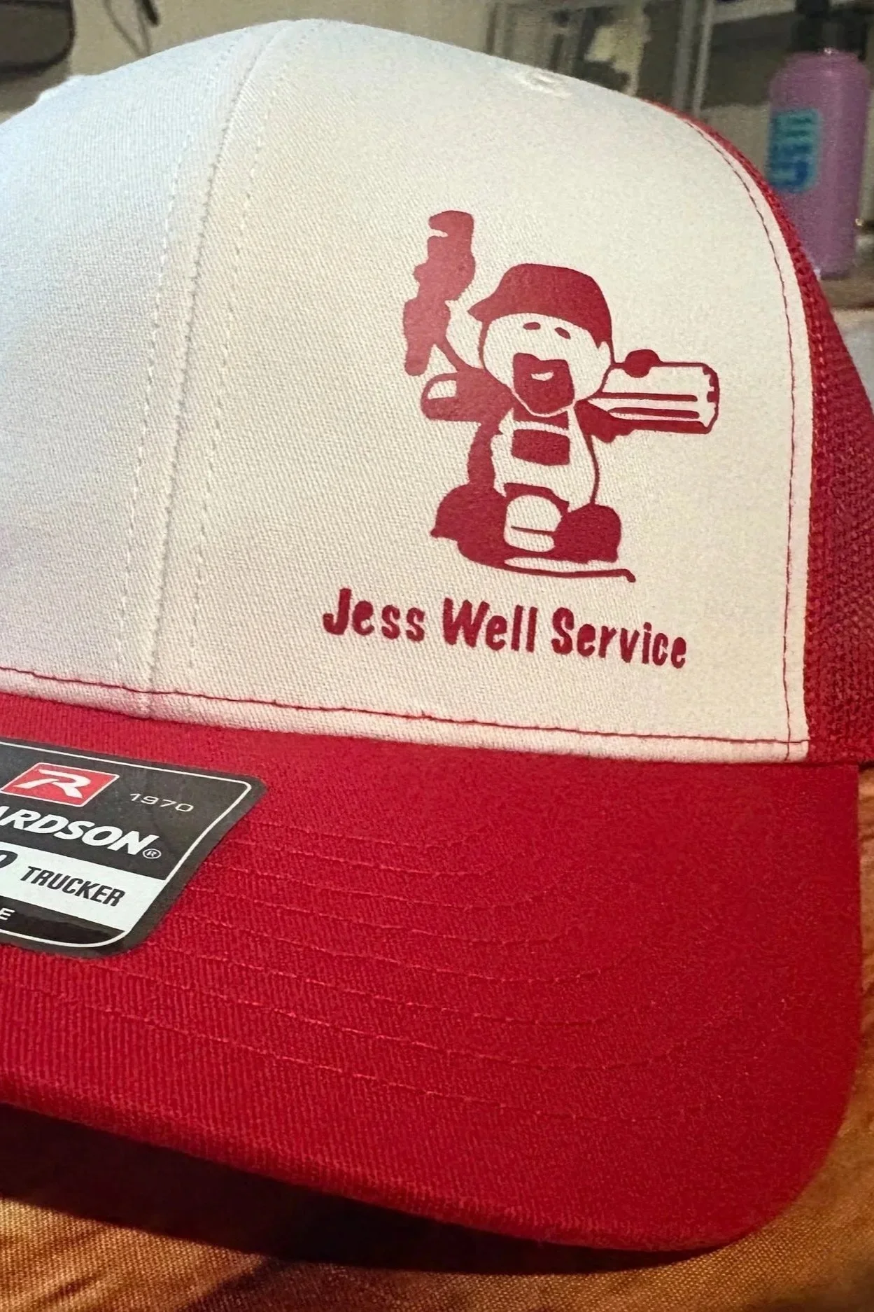A red and white trucker hat with a cartoon character logo and the text 'Jess Well Service' embroidered on the front. The character is holding a tool and wearing a cap.