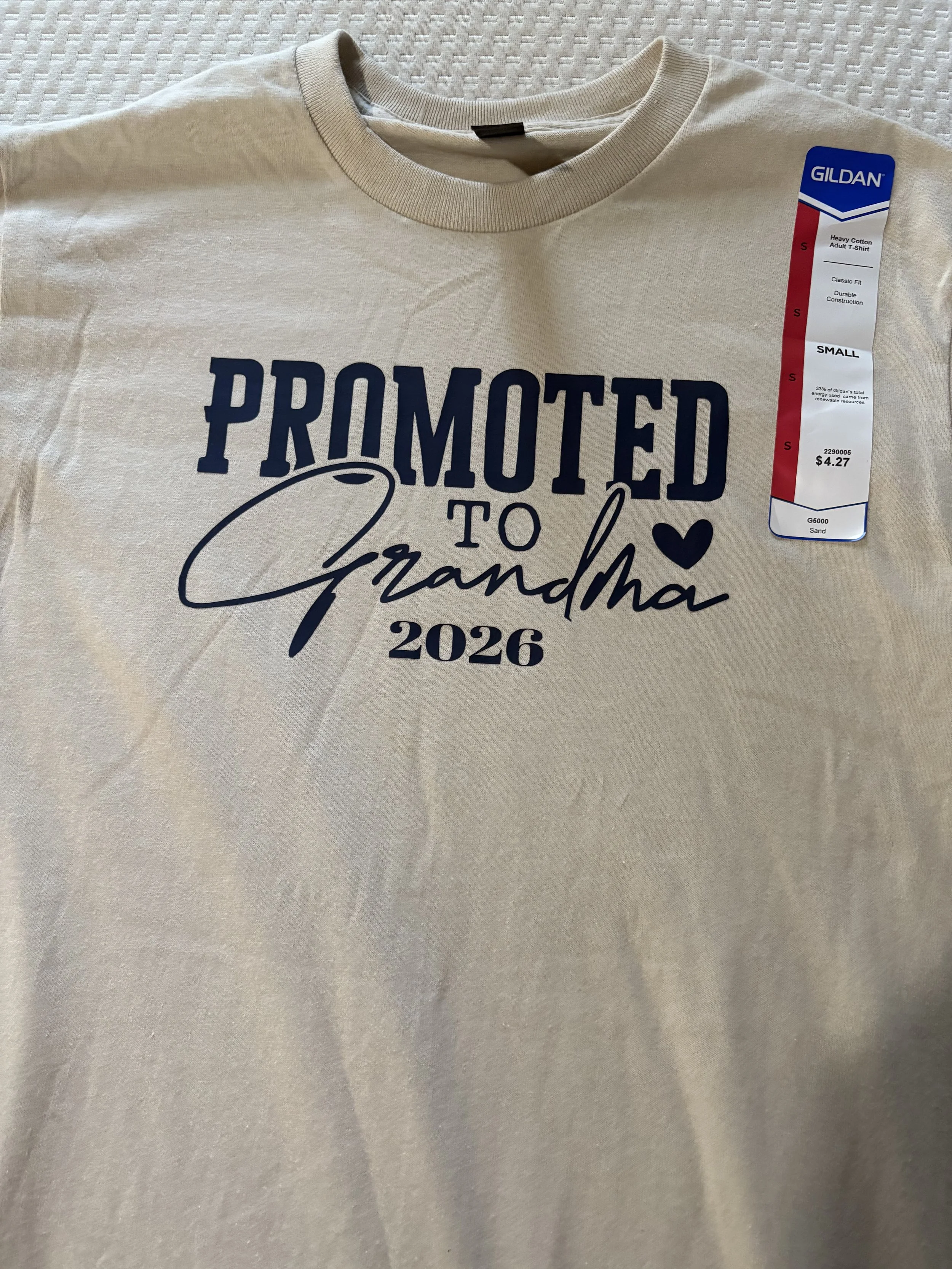 A cream-colored T-shirt with black and white graphics, including the text 'PROMOTED TO Grandma 2026' and a signature-style name, laid out on a textured surface, with a Gildan brand tag attached.