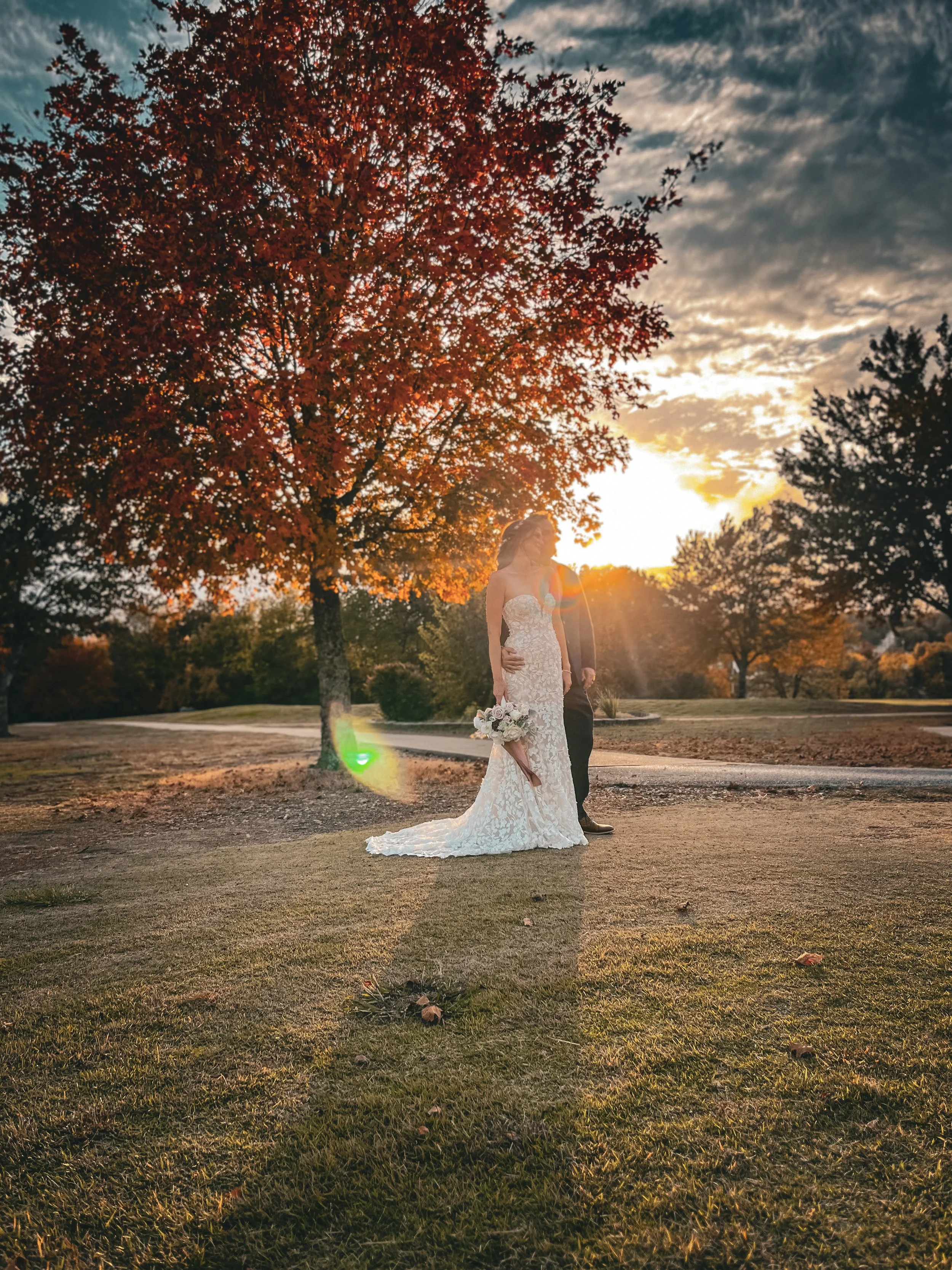 A bride in a white wedding dress holding a bouquet standing on grass during sunset, with a large tree with red leaves and a partly cloudy sky in the background.