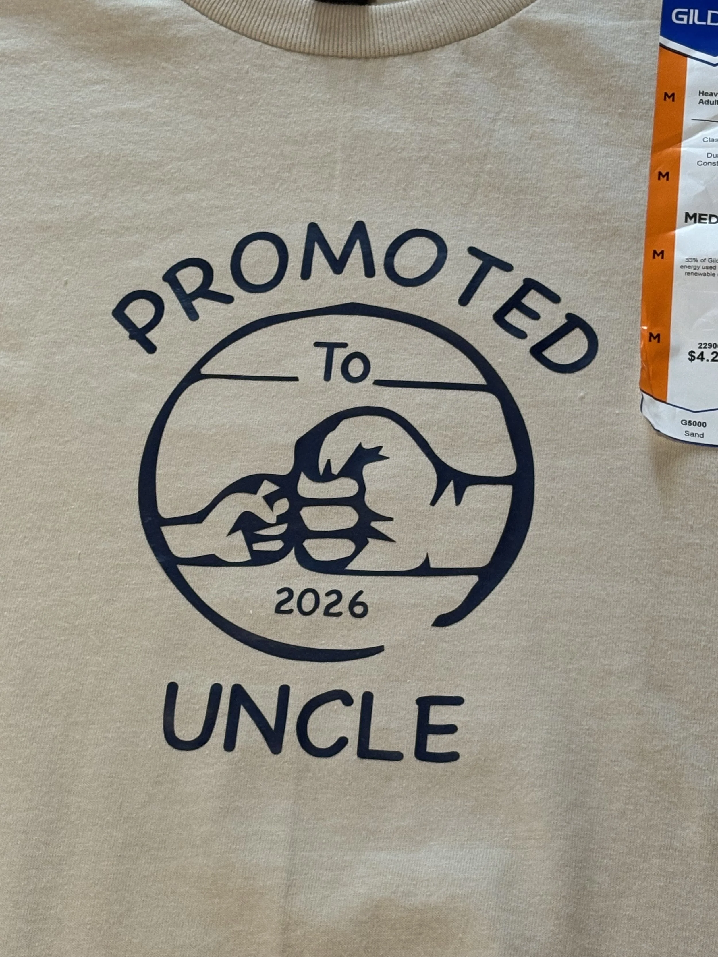 A beige T-shirt with a graphic design including a circular emblem featuring a dog and the words 'Promoted to Uncle 2026' and part of a bag or package of Gildan brand sock or stocking lying partially visible on the right side.